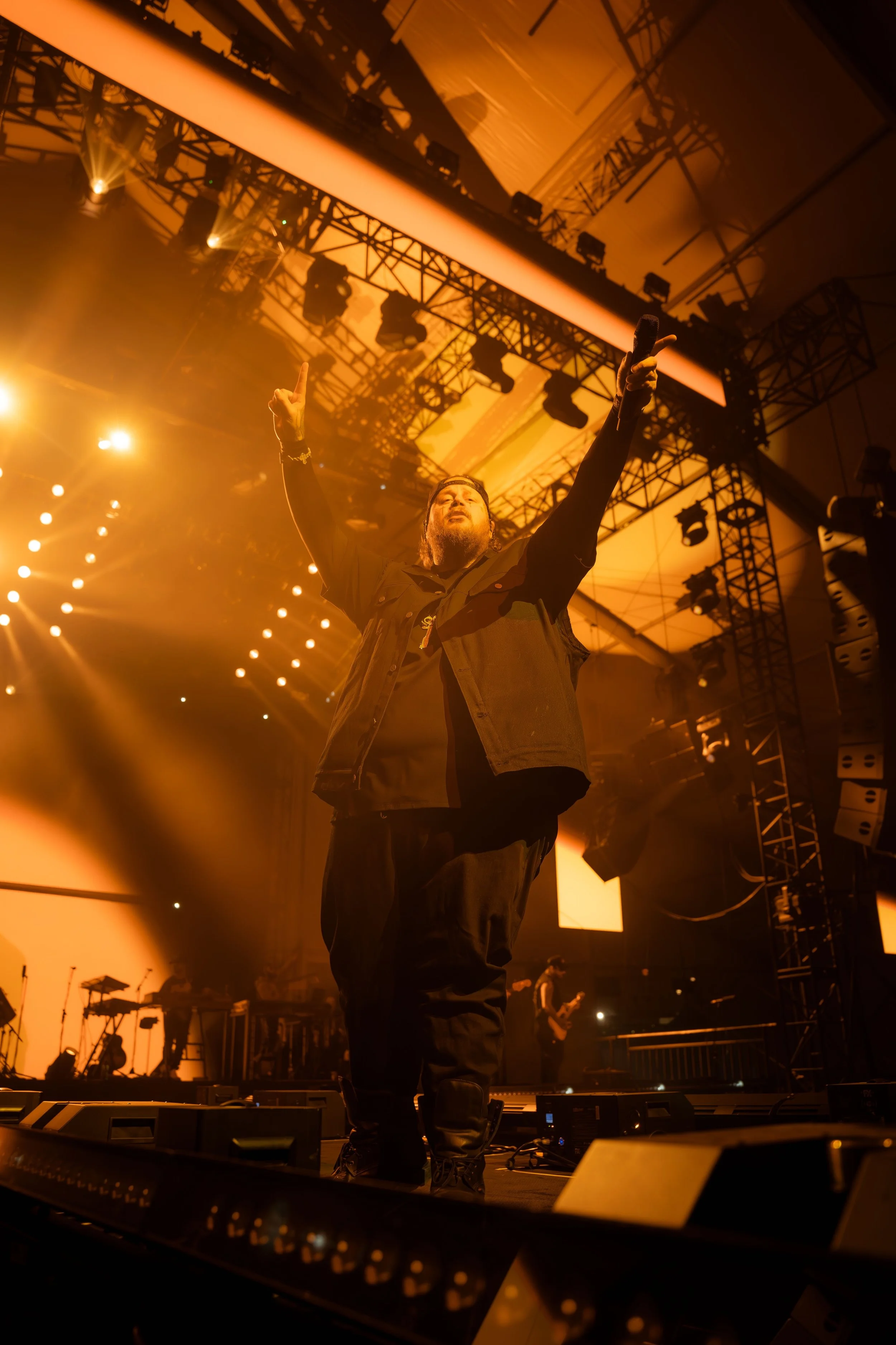 Male performer on stage at concert with arms raised, surrounded by stage lights and equipment.