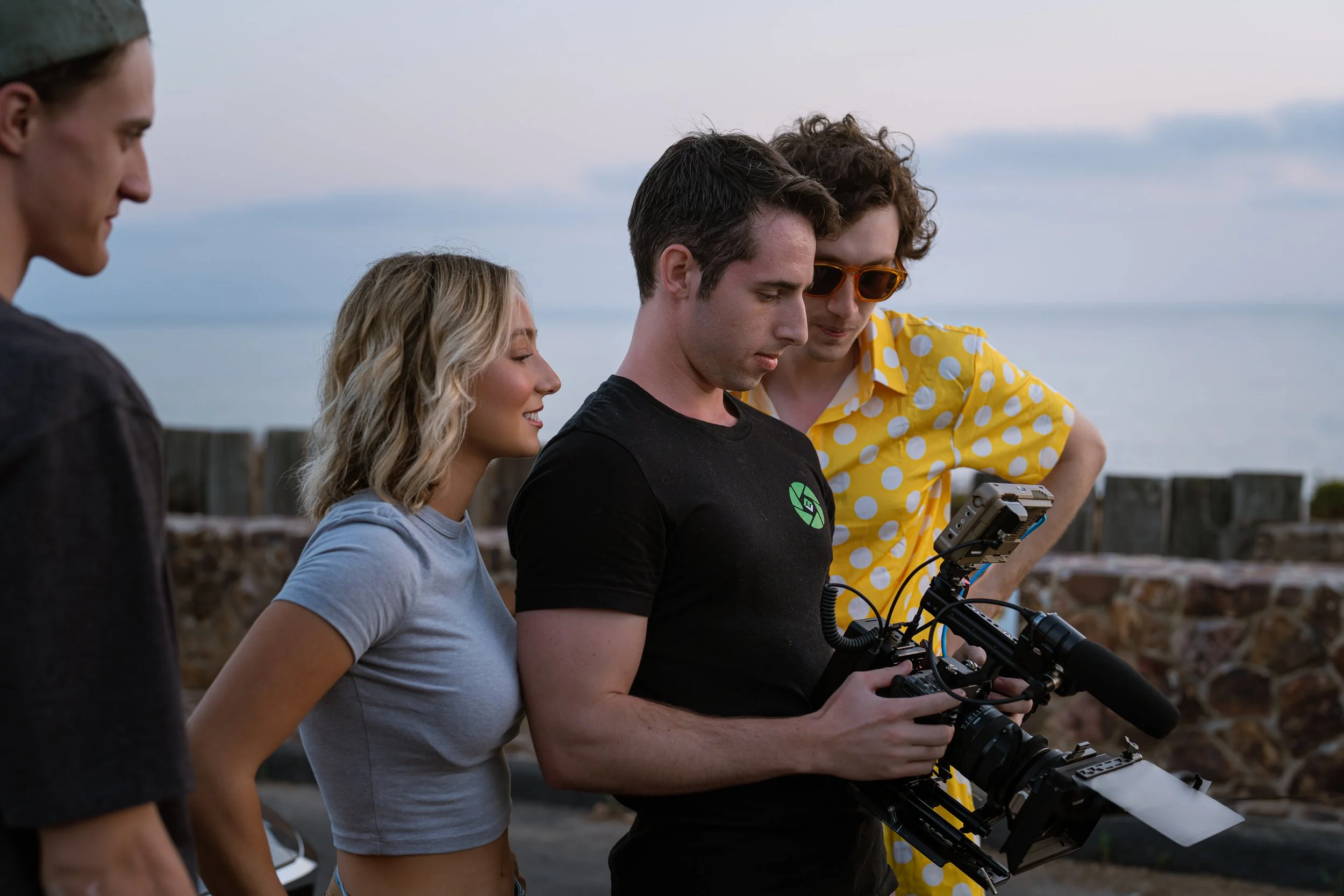 Group of four young people, one holding a camera with a mounted monitor, standing near a stone wall by the water during sunset.