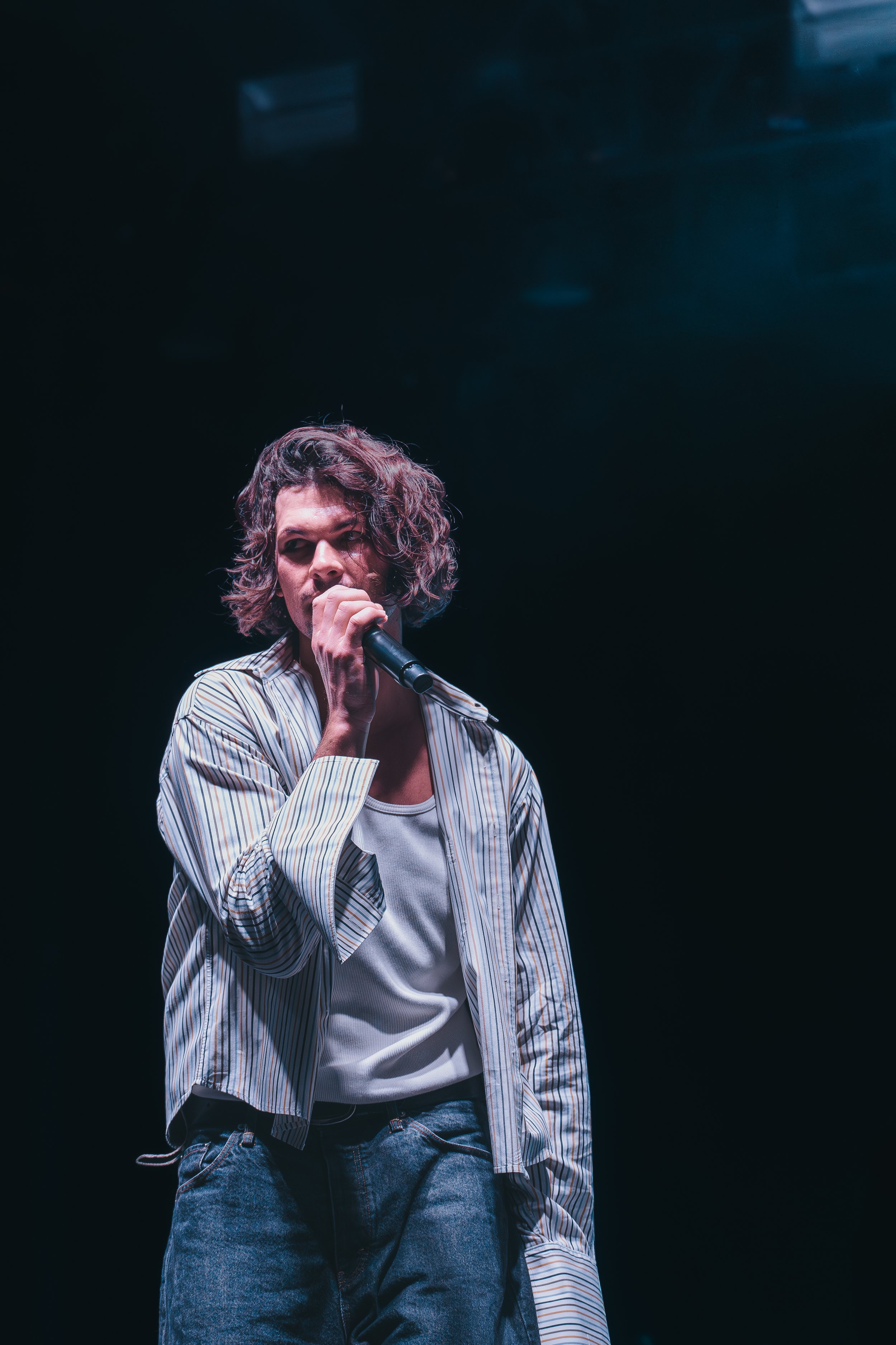 A person with curly hair holding a microphone on a dark stage.