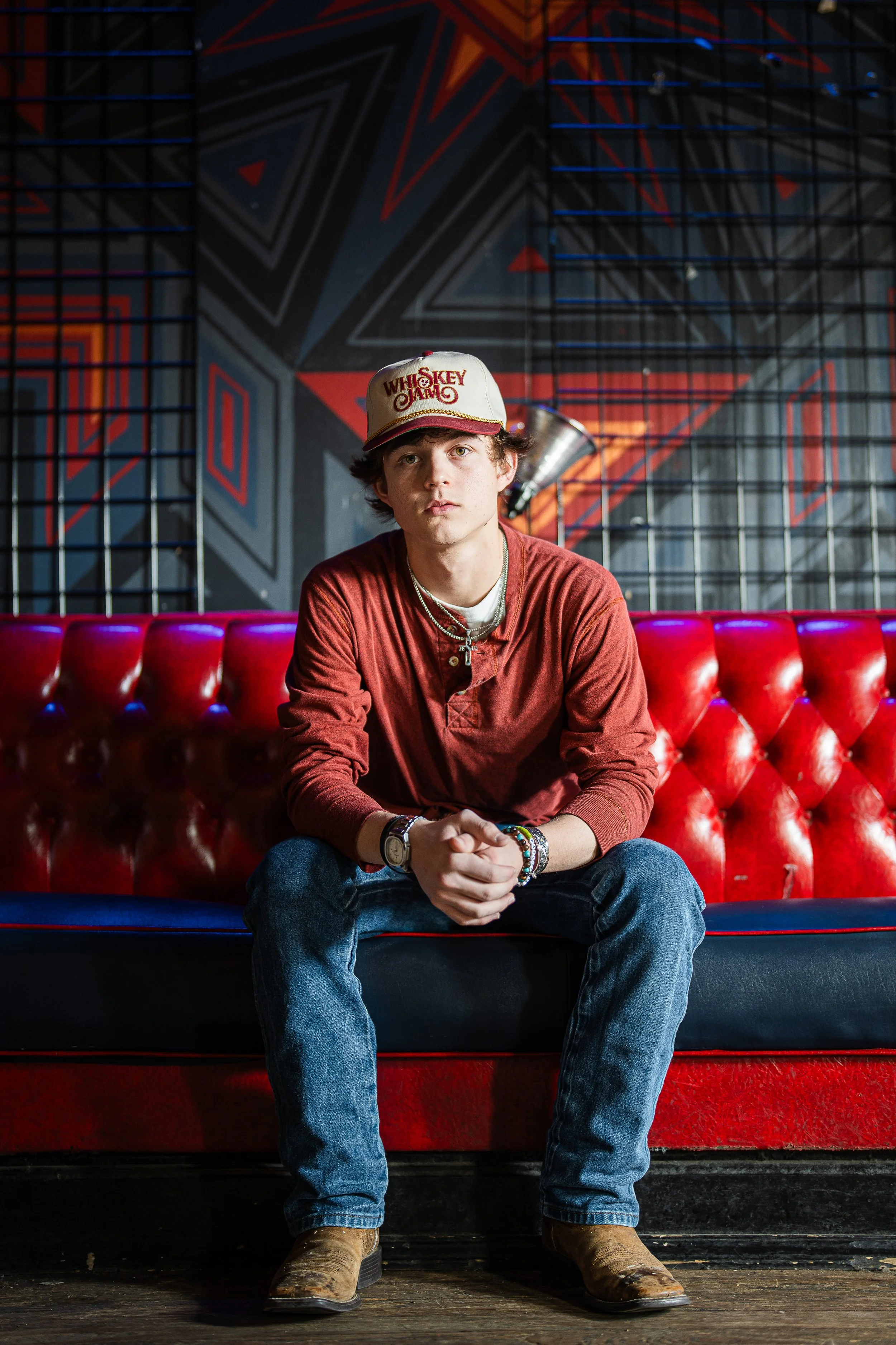 A young man with brown hair sitting on a red leather bench in a room with a colorful geometric mural, wearing a beige baseball cap with red text, a red long-sleeve shirt, blue jeans, and brown boots.
