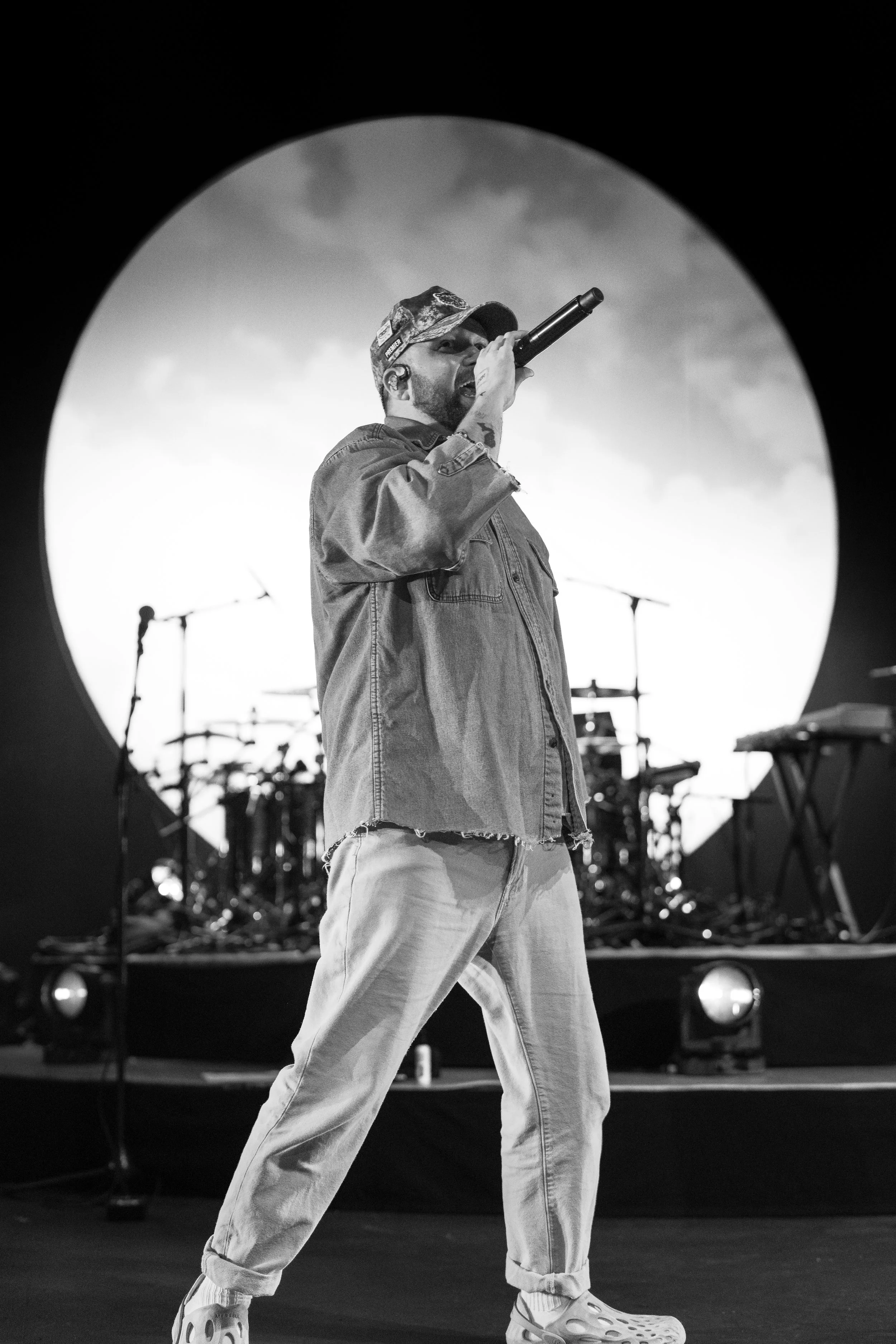 A male singer performing on stage with a microphone, wearing a cap, denim jacket, and sneakers, with drums and keyboard in the background, and a large circular backdrop.