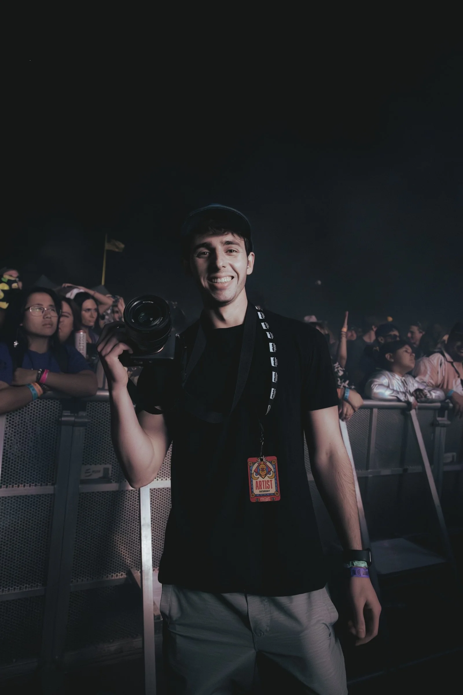 A young man at a concert or music event holding a camera, wearing an artist pass around his neck, with a crowd of people behind a barricade.