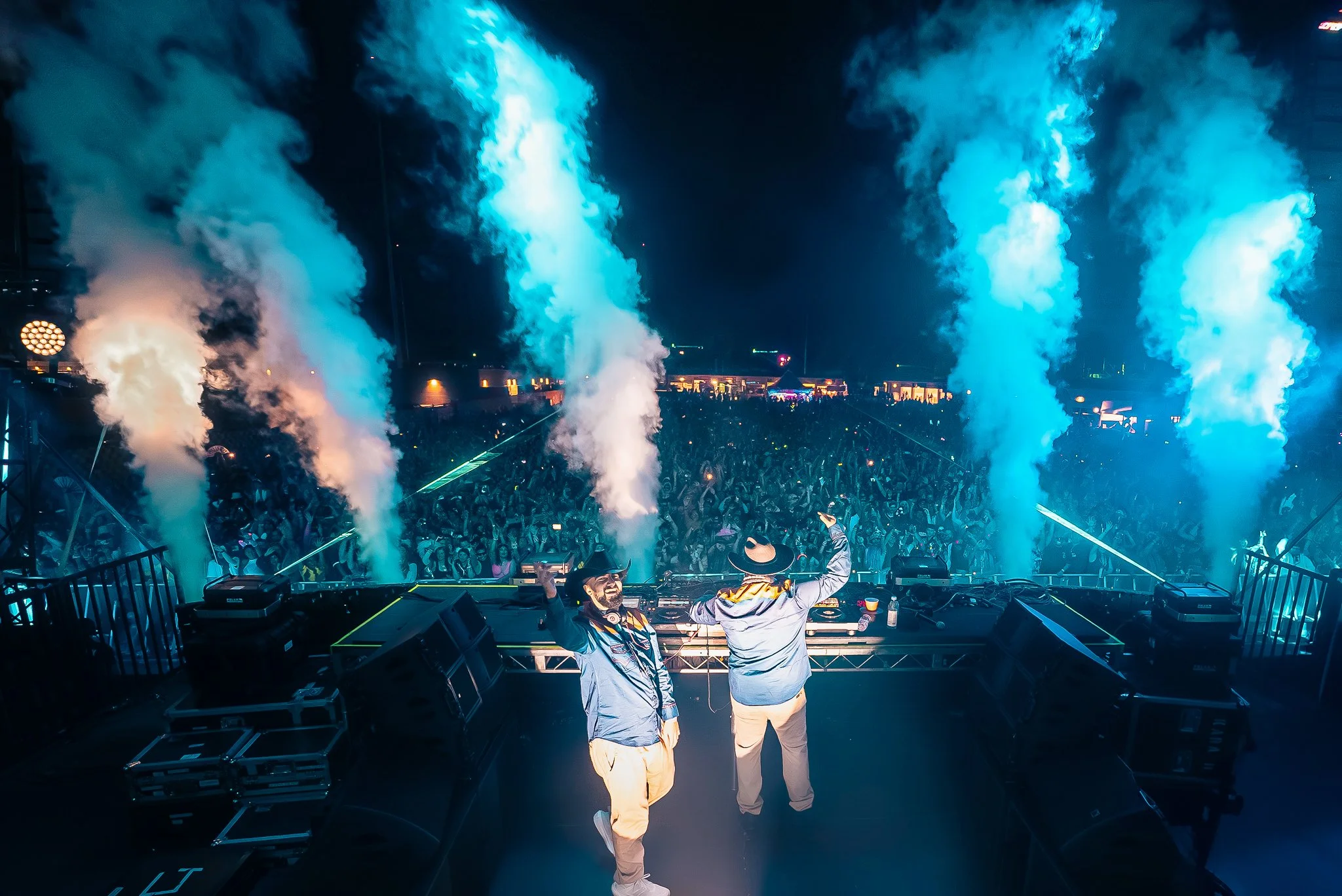 Two DJs on stage at night with large crowd, colorful smoke effects, and bright lighting at a concert or festival.
