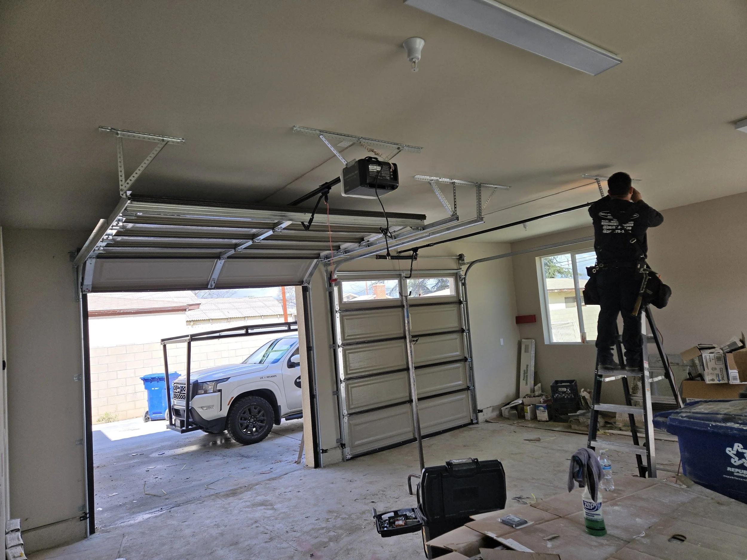 A worker standing on a ladder installing or repairing a garage door opener system inside a garage. The garage door is partially open, revealing a parked vehicle and a blue recycling bin outside. Various tools and equipment are scattered around.