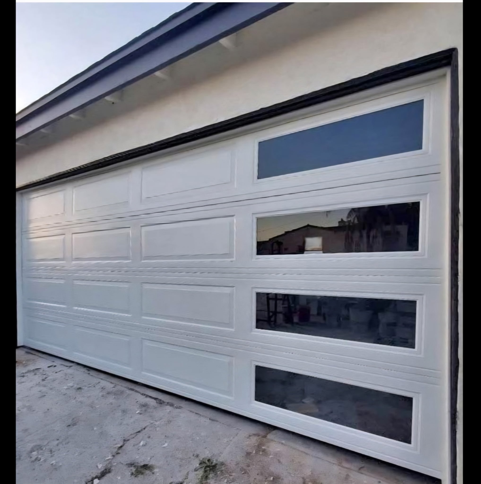 A white sectional garage door with four horizontal windows at the top and bottom, installed on a house exterior with a concrete driveway in front.