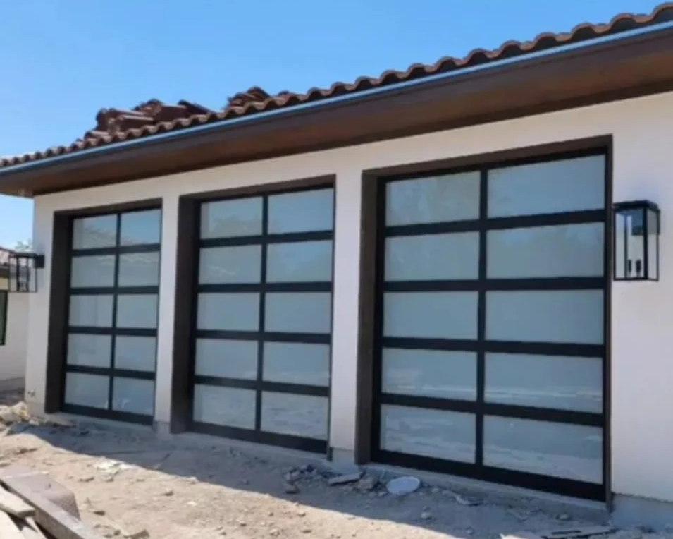 Three modern garage doors with glass panels on a house under construction, with a light fixture on the wall and a clear blue sky above.