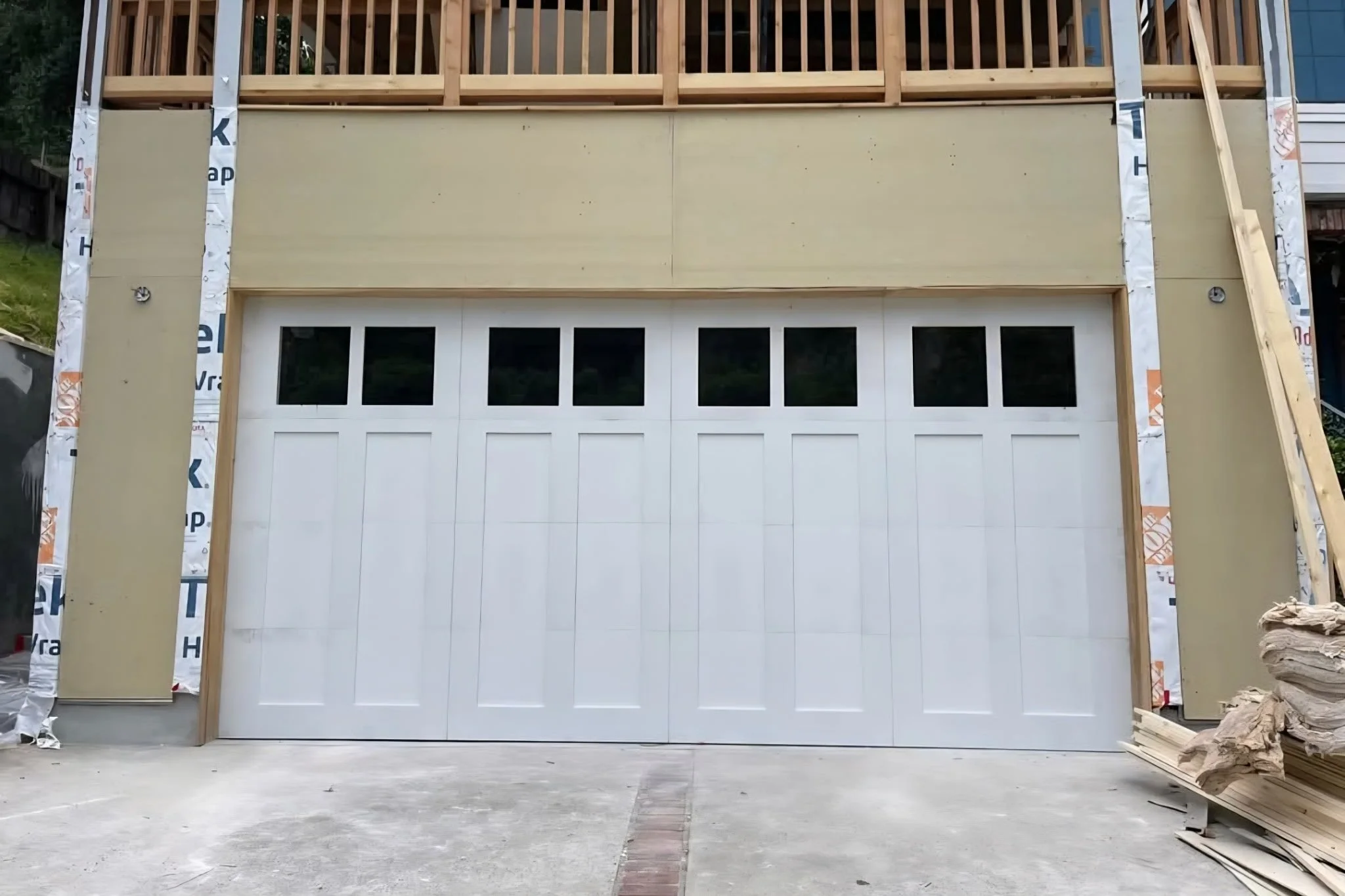 The image shows a garage under construction with a white garage door and a second-story framework with wooden beams. There is construction material around the site.