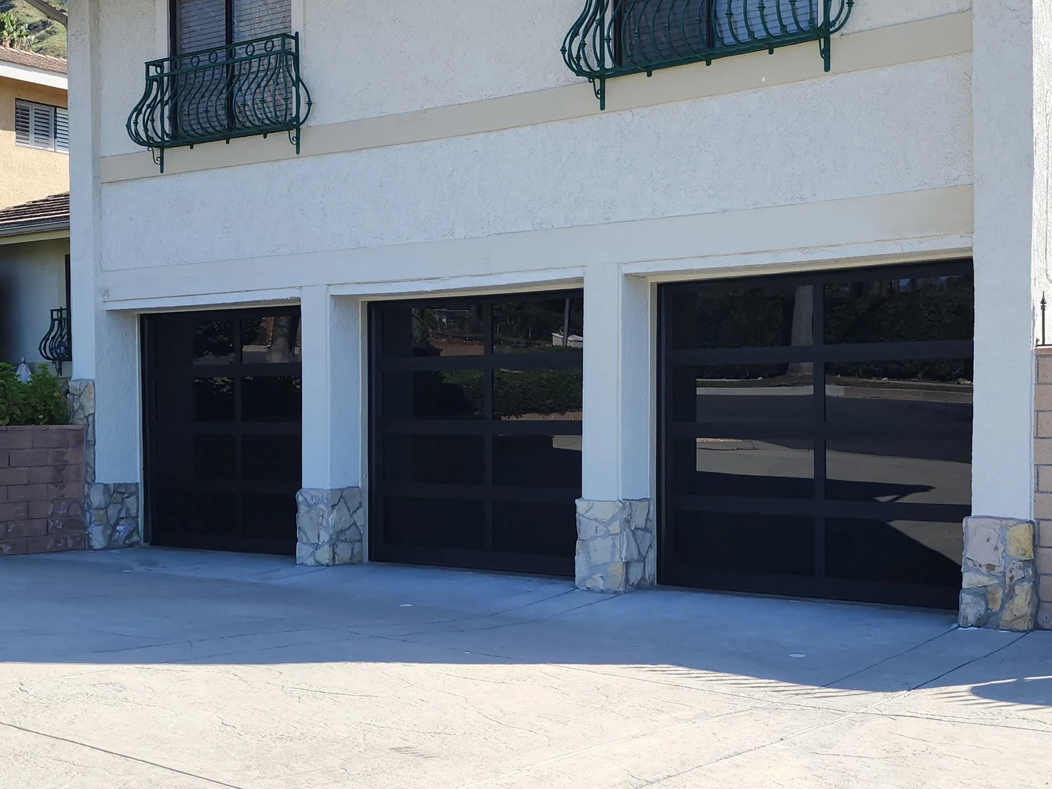 Three black garage doors with stone accents at the bottom, part of a building with white textured walls and green balcony railings.