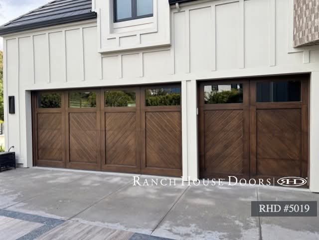 A house with a large wooden garage door and a small window above it. The house exterior is painted white with vertical siding. There is a concrete driveway in front of the garage.