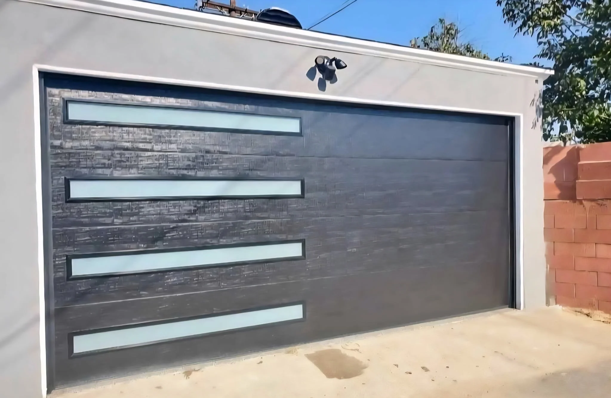 Modern garage door with horizontal windows and textured black panels, surrounded by a white wall and brick wall on the right, under a clear blue sky.