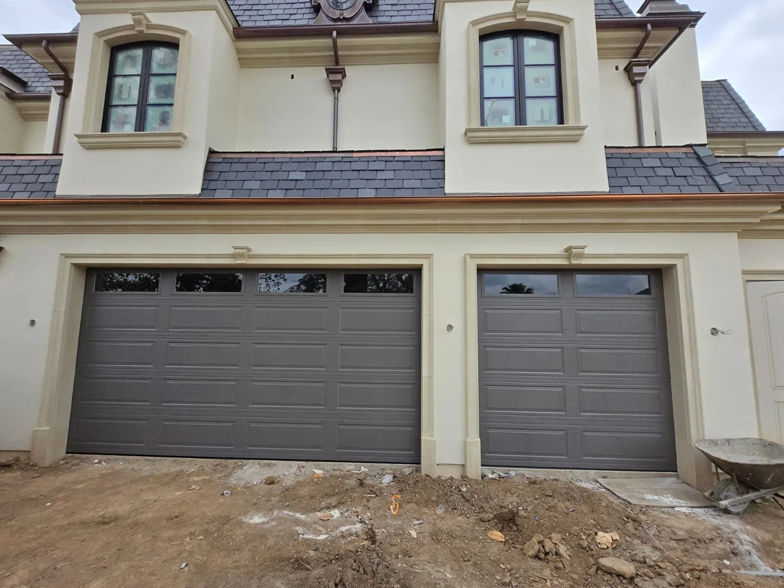 Exterior view of a modern house with two garage doors. The house has beige walls, decorative window frames, and a dark shingled roof. The ground in front is under construction with dirt and rocks, and a wheelbarrow is visible on the right side.