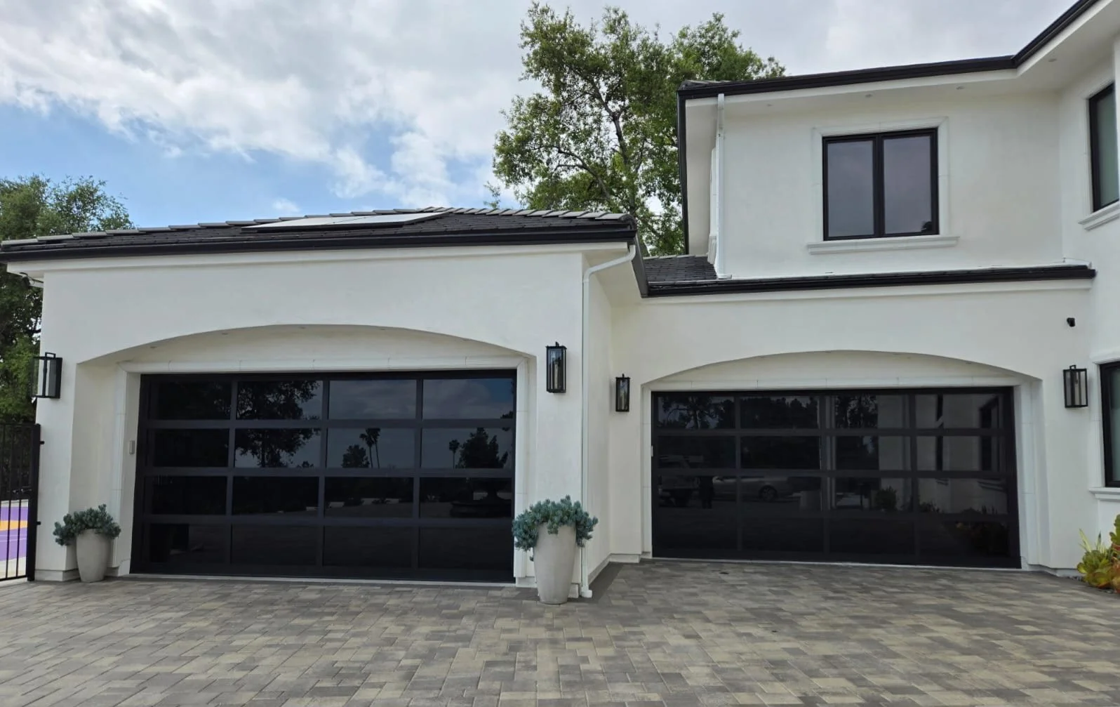 Modern white house with two black garage doors, potted plants on either side, and exterior wall lights. Paved driveway in front, trees in the background, and cloudy sky.