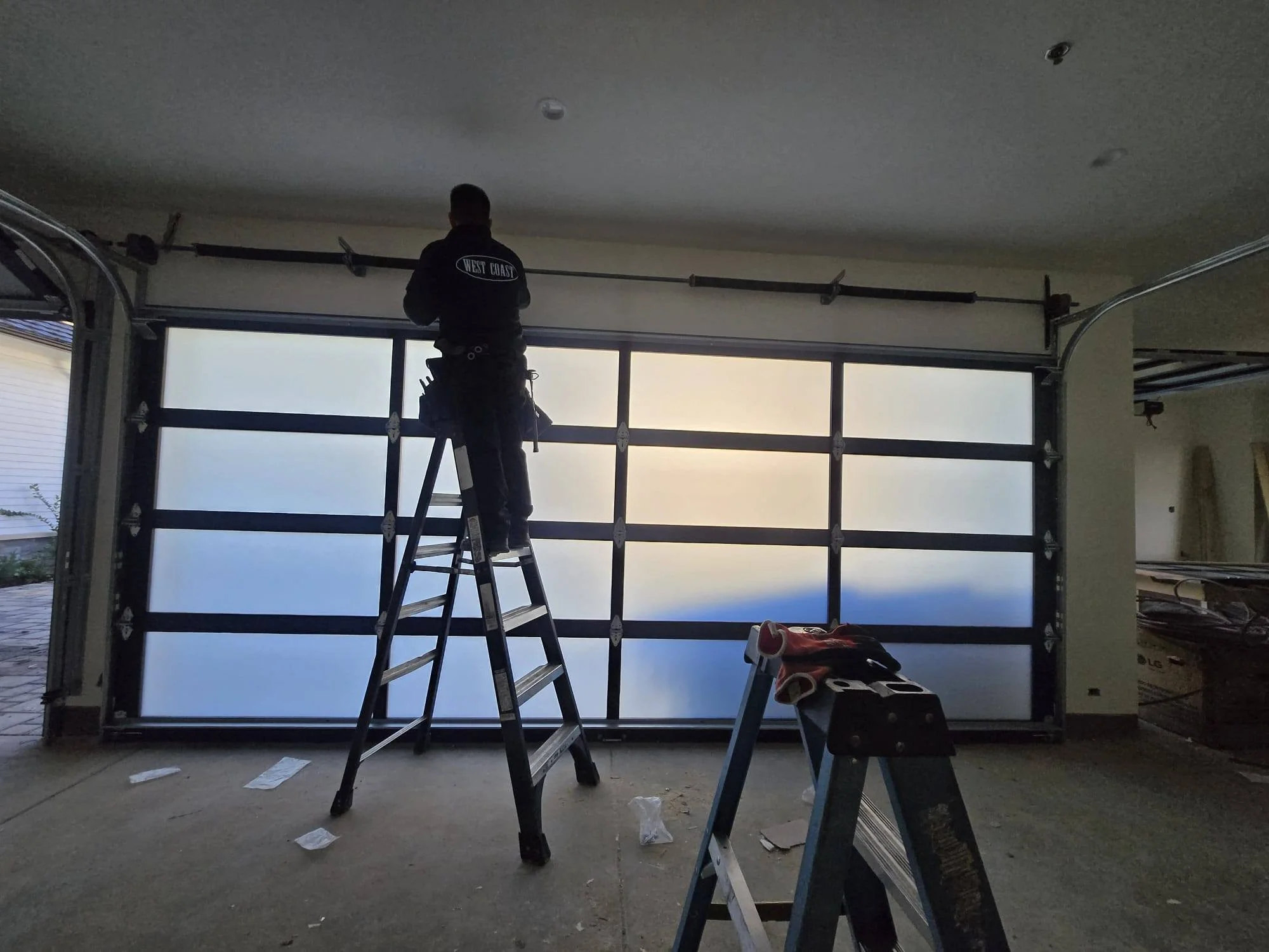 A worker standing on a ladder installing or repairing a large garage door with frosted glass panels inside a garage.