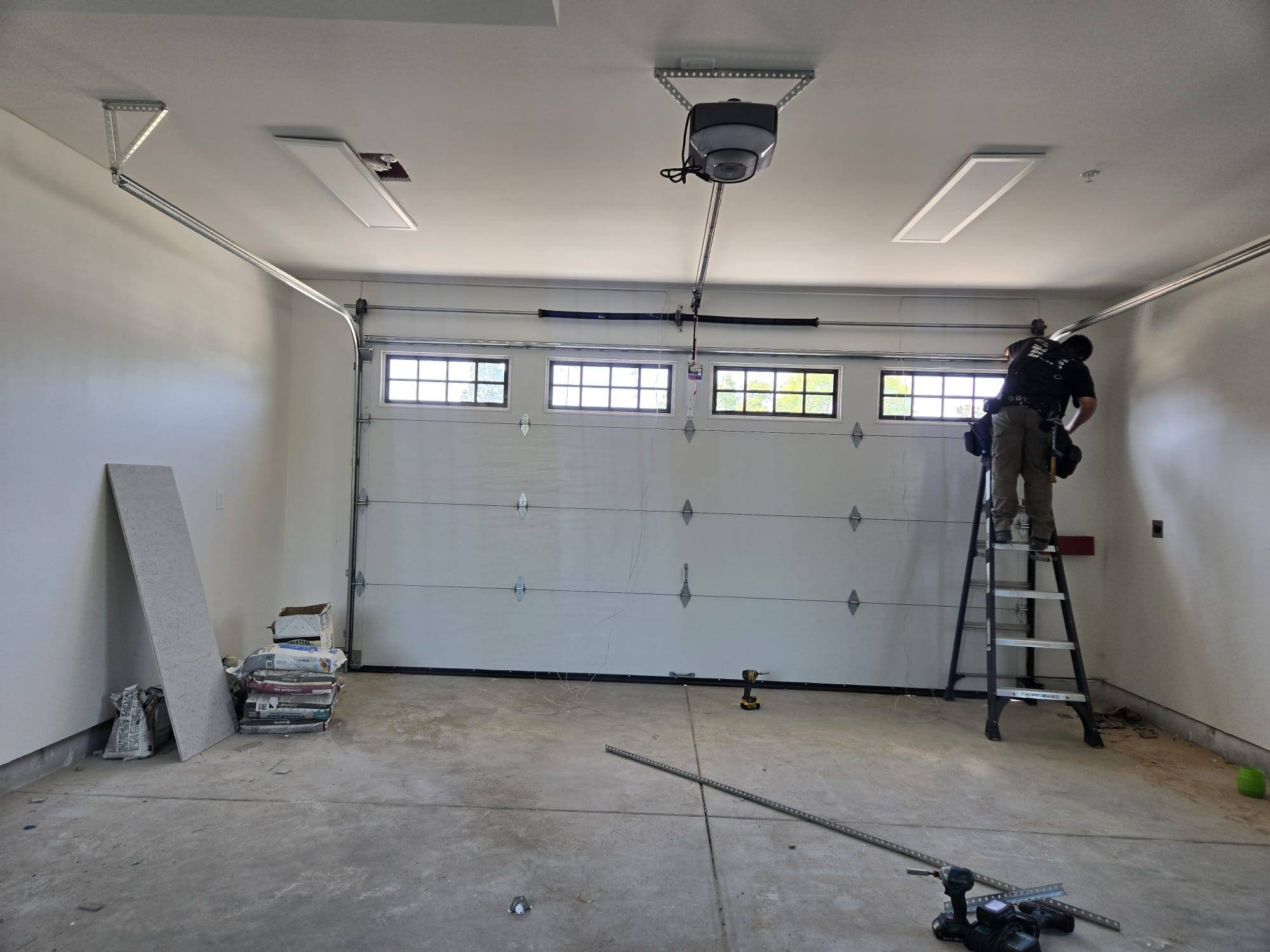 A person standing on a ladder working on the garage door opener in a garage under construction.