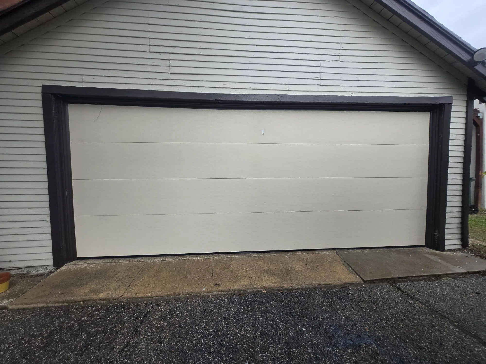 An automatic garage door with a black frame on a beige background, part of a house with white siding and a sloped roof.