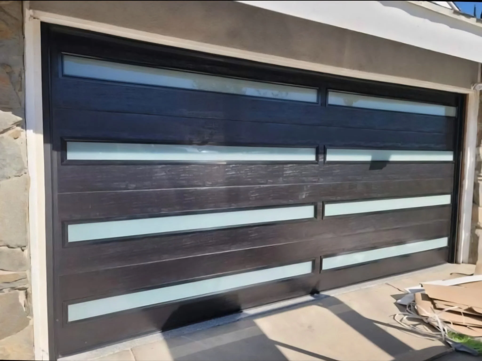 A modern garage door with horizontal dark wooden panels and narrow horizontal frosted glass windows, set within a stone and concrete exterior wall.