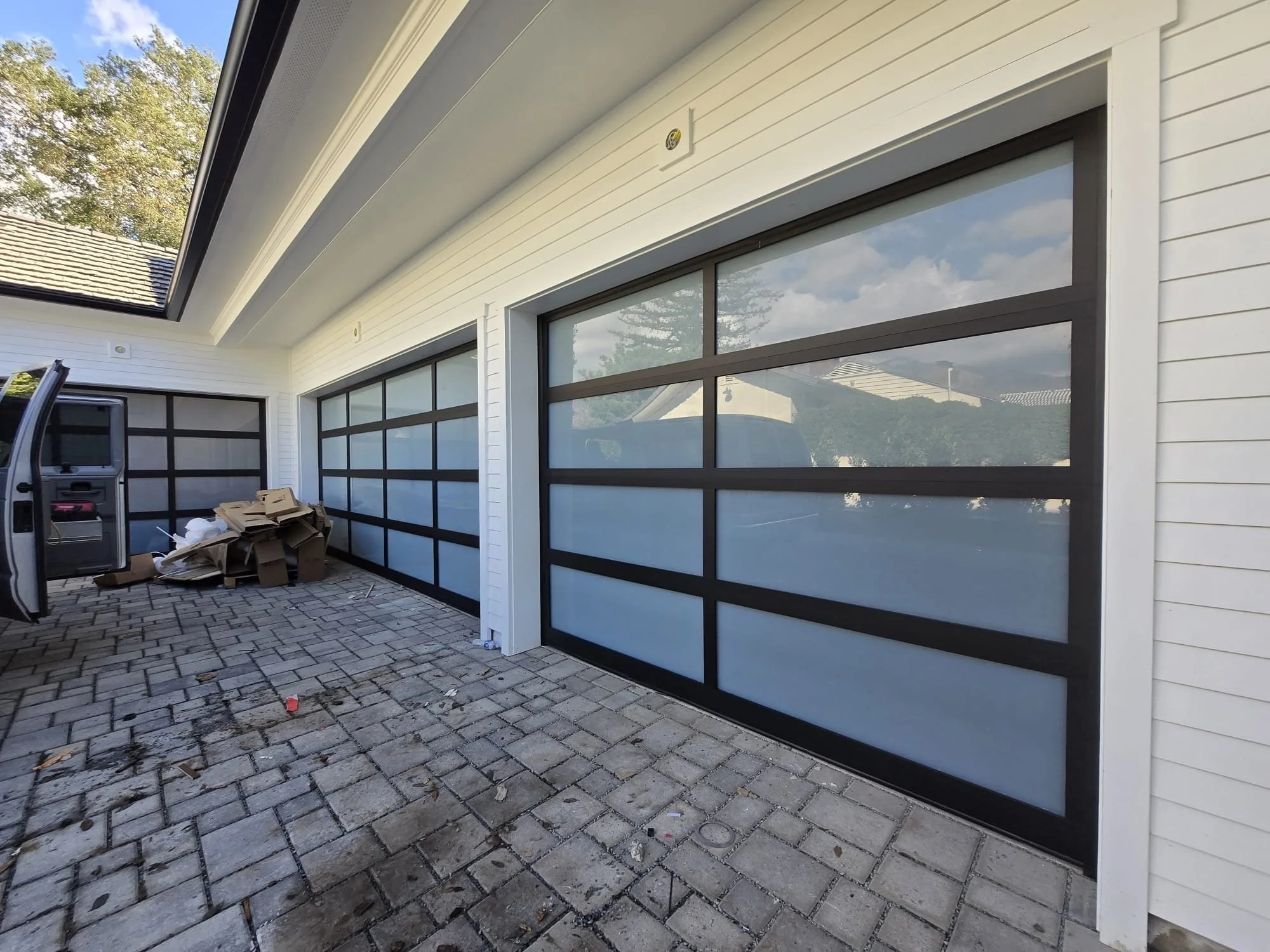 View of a garage with large glass doors, white siding, and a paved driveway under a clear sky.