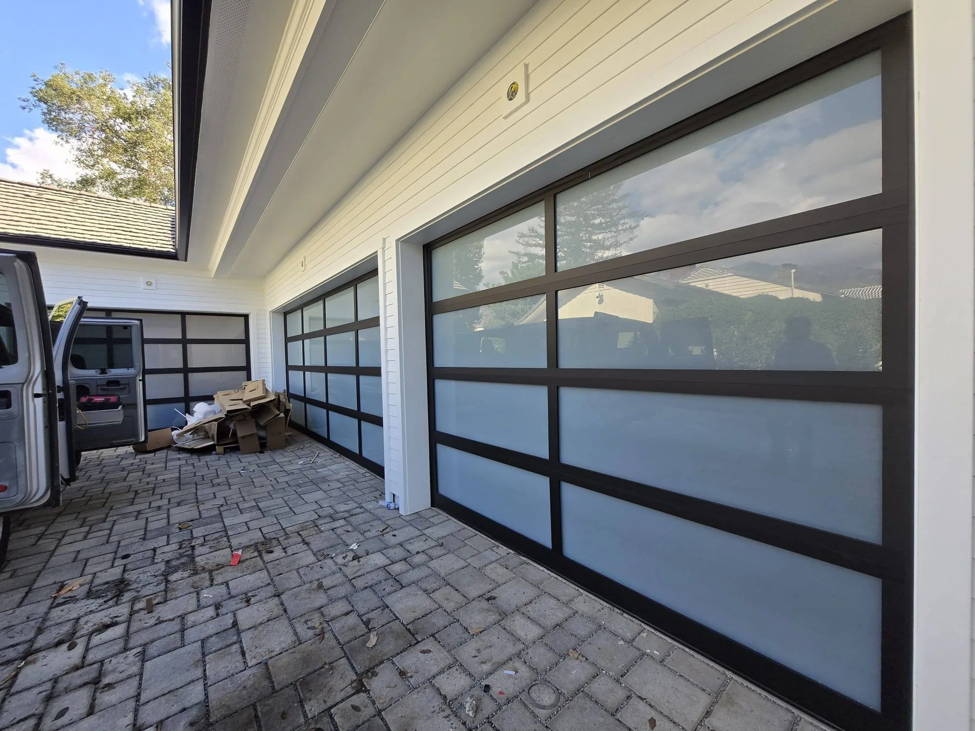 Residential garage with black-framed frosted glass doors, driveway with pavers, and a van parked nearby.