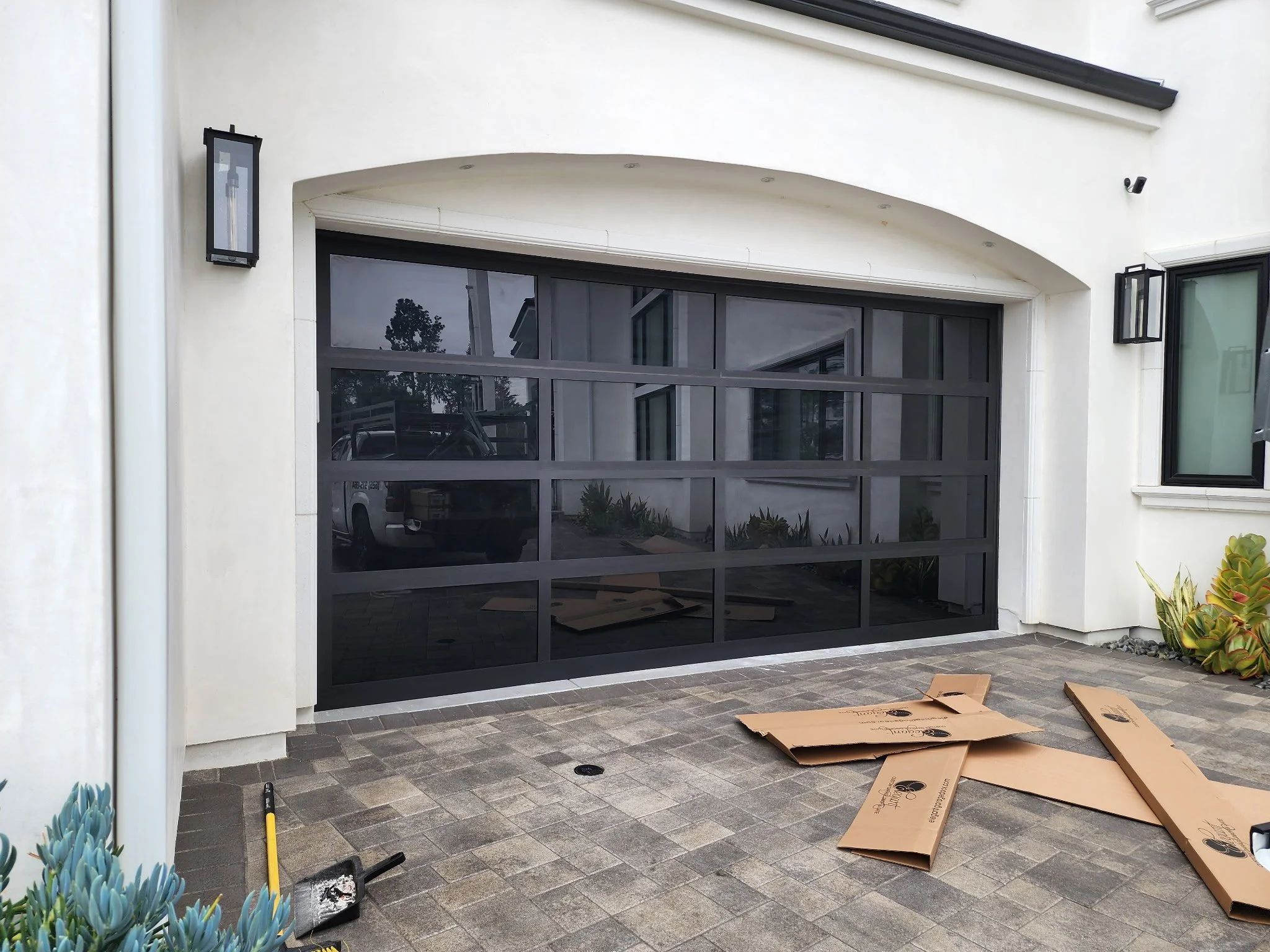A modern house garage with a black glass garage door, outside patio with paving stones, and packaged cardboard boxes on the ground. Wall-mounted outdoor light fixtures on either side of the garage door. Reflection of trees and a parked white vehicle seen in the garage door window.