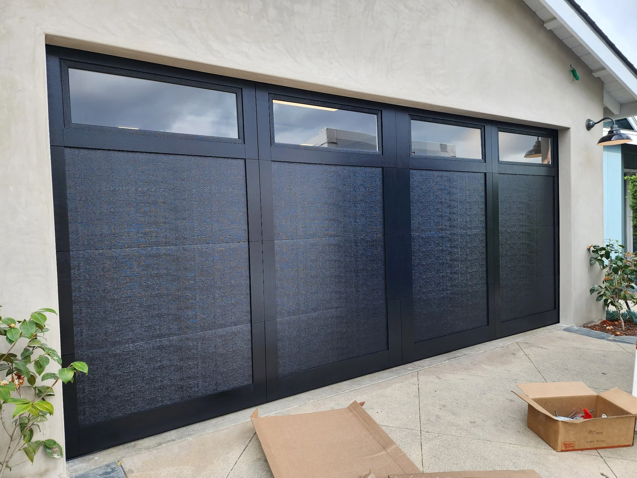 Large black garage door with small upper windows, located in a modern house with a beige exterior wall and small plants on either side.