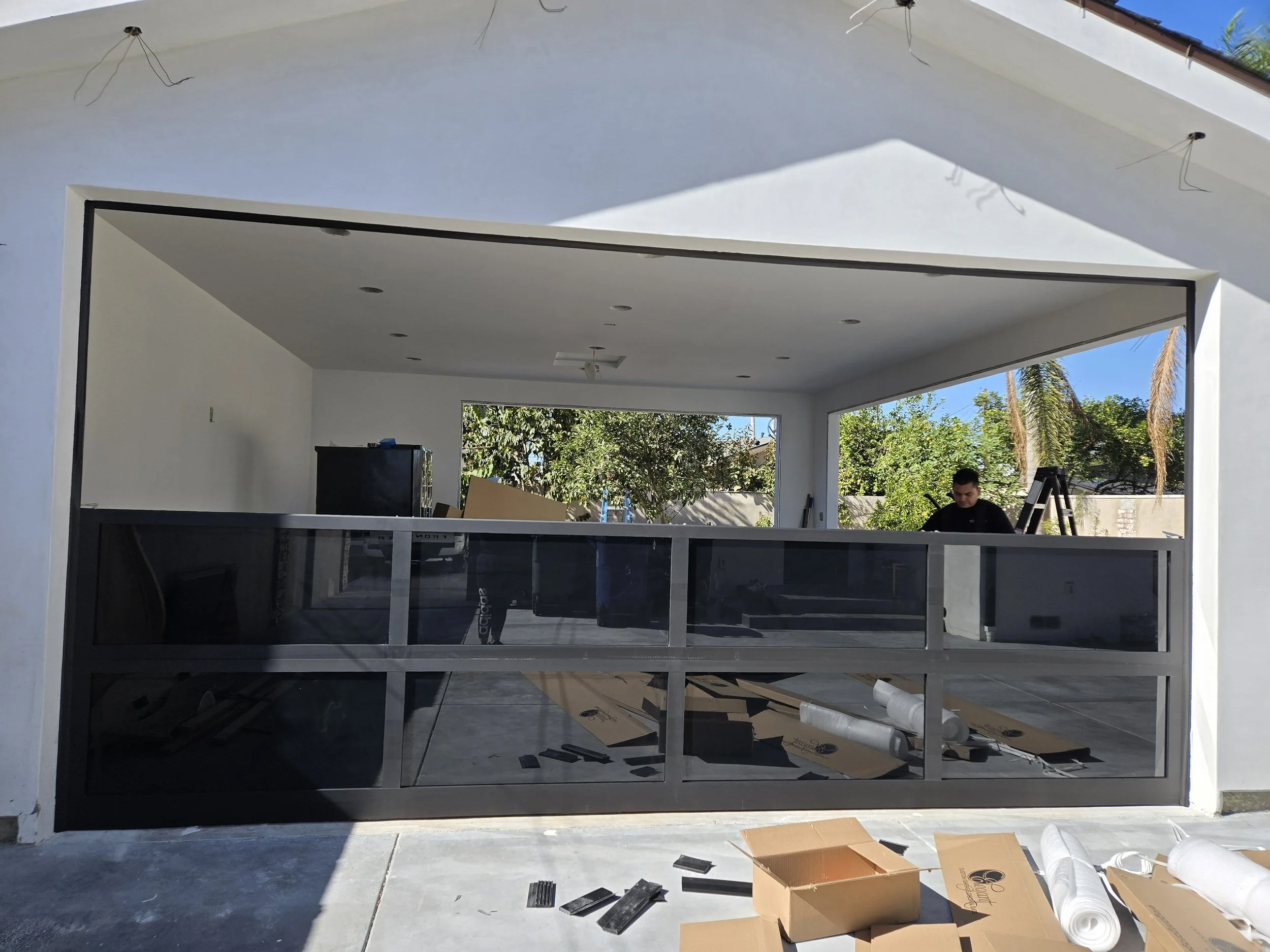 Construction workers installing a glass garage door in a modern house with white walls and an open interior view, surrounded by trees and blue sky.