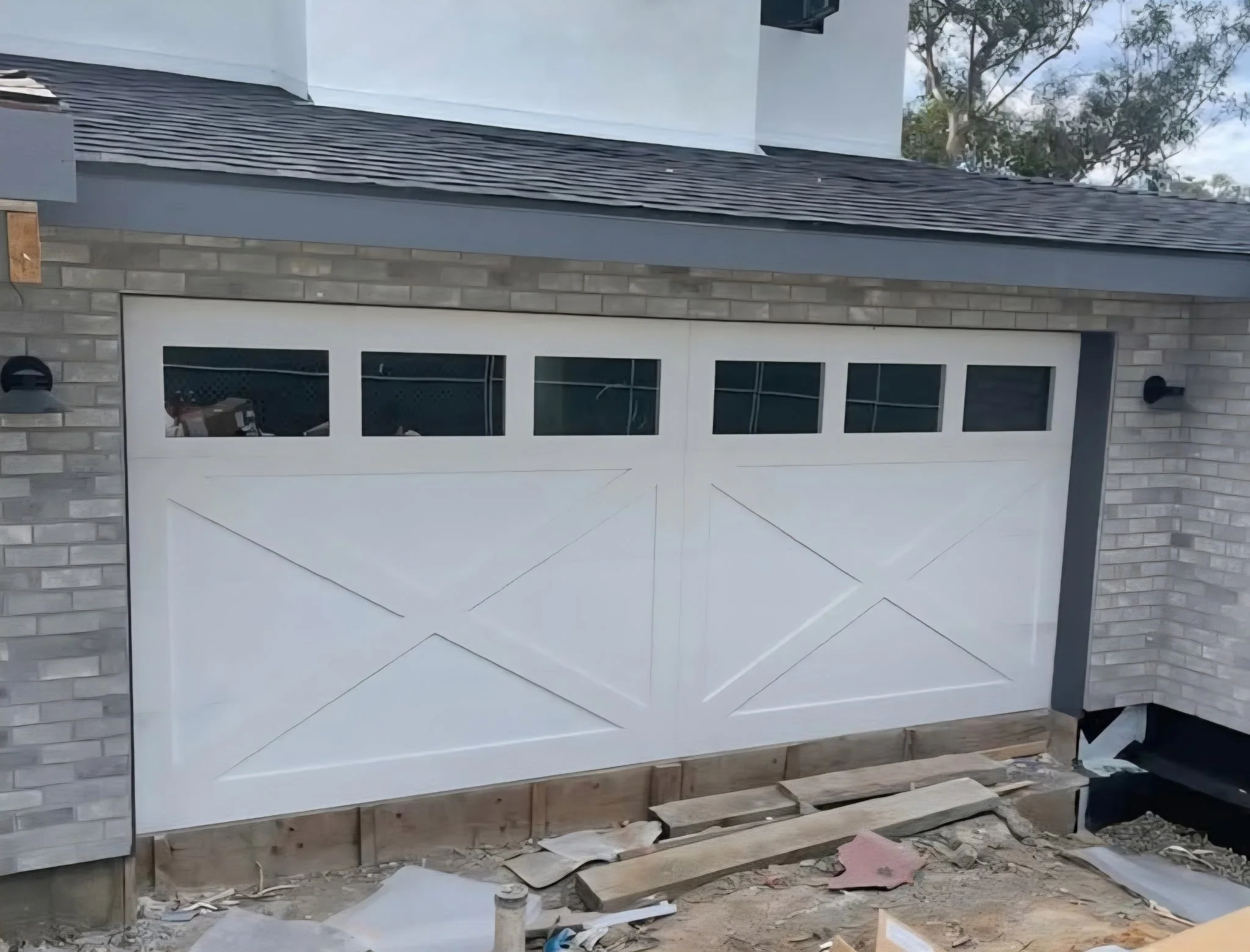 View of a white garage door with windows at the top, surrounded by gray brick wall, with construction materials and debris in front and trees visible in the background.