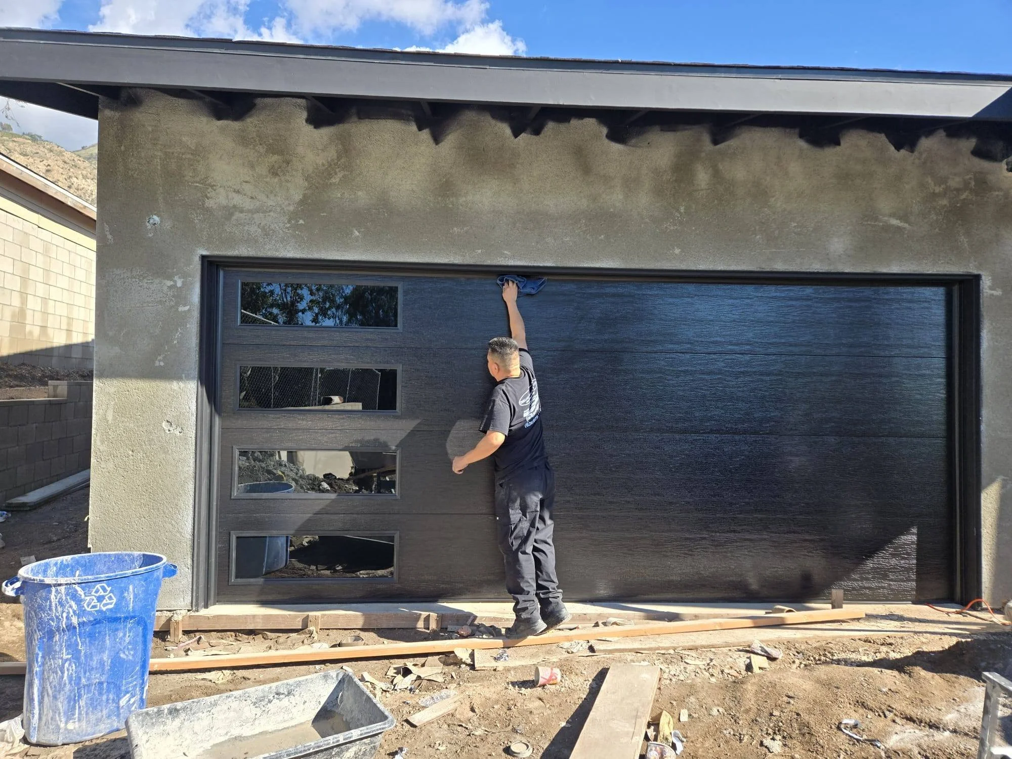 A person installs a dark gray garage door with four small rectangular windows on the left side, outside a building under construction. Construction tools and debris are on the ground.