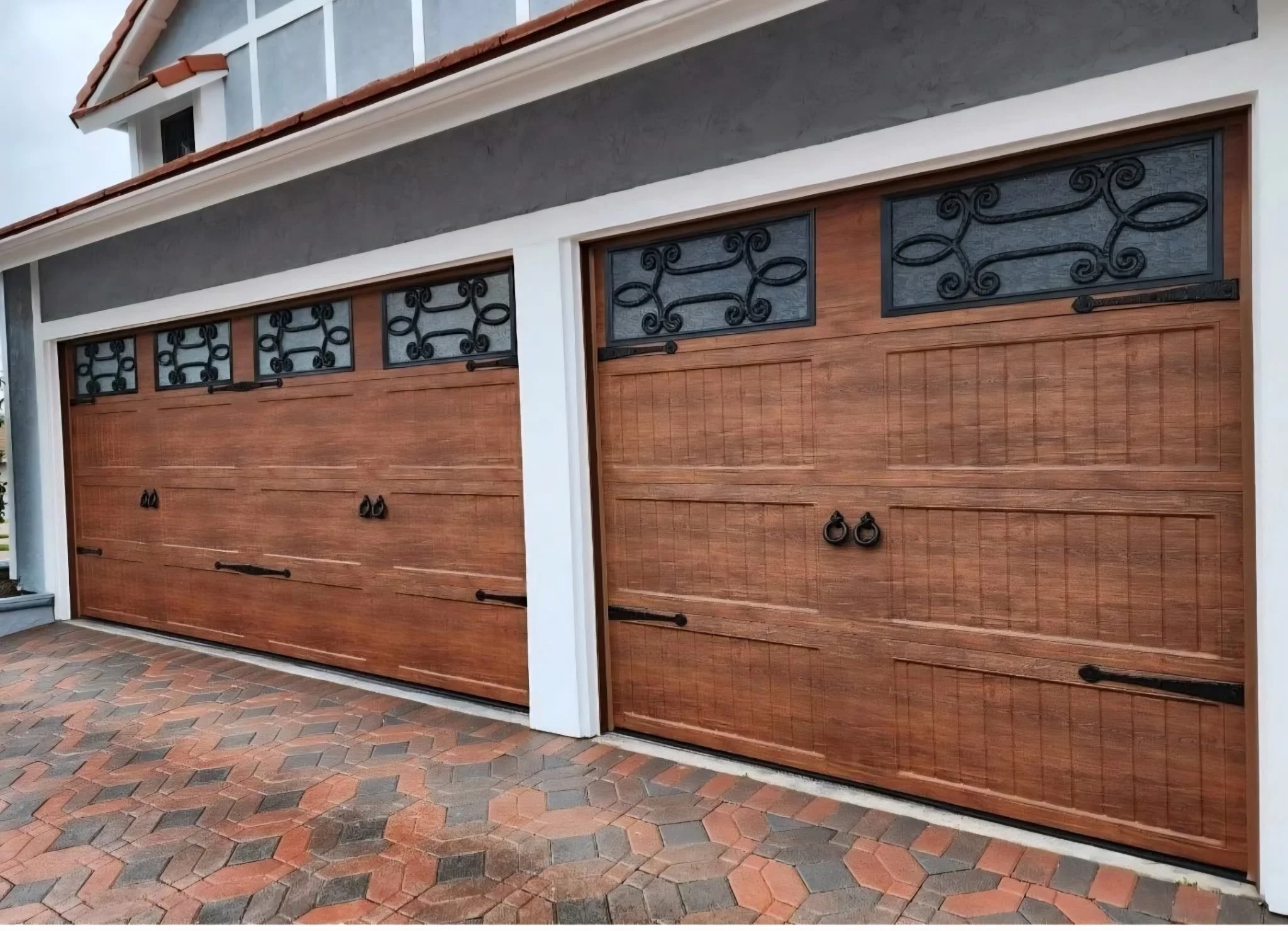 Two wooden garage doors with black decorative wrought iron windows and handles, set in a modern house with gray walls and a brick-paved driveway.