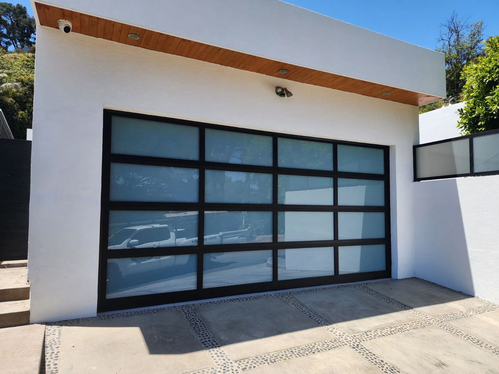 Modern house with a white exterior, large glass garage door, and a wooden accent ceiling above the entrance, set against a background of trees and clear blue sky.