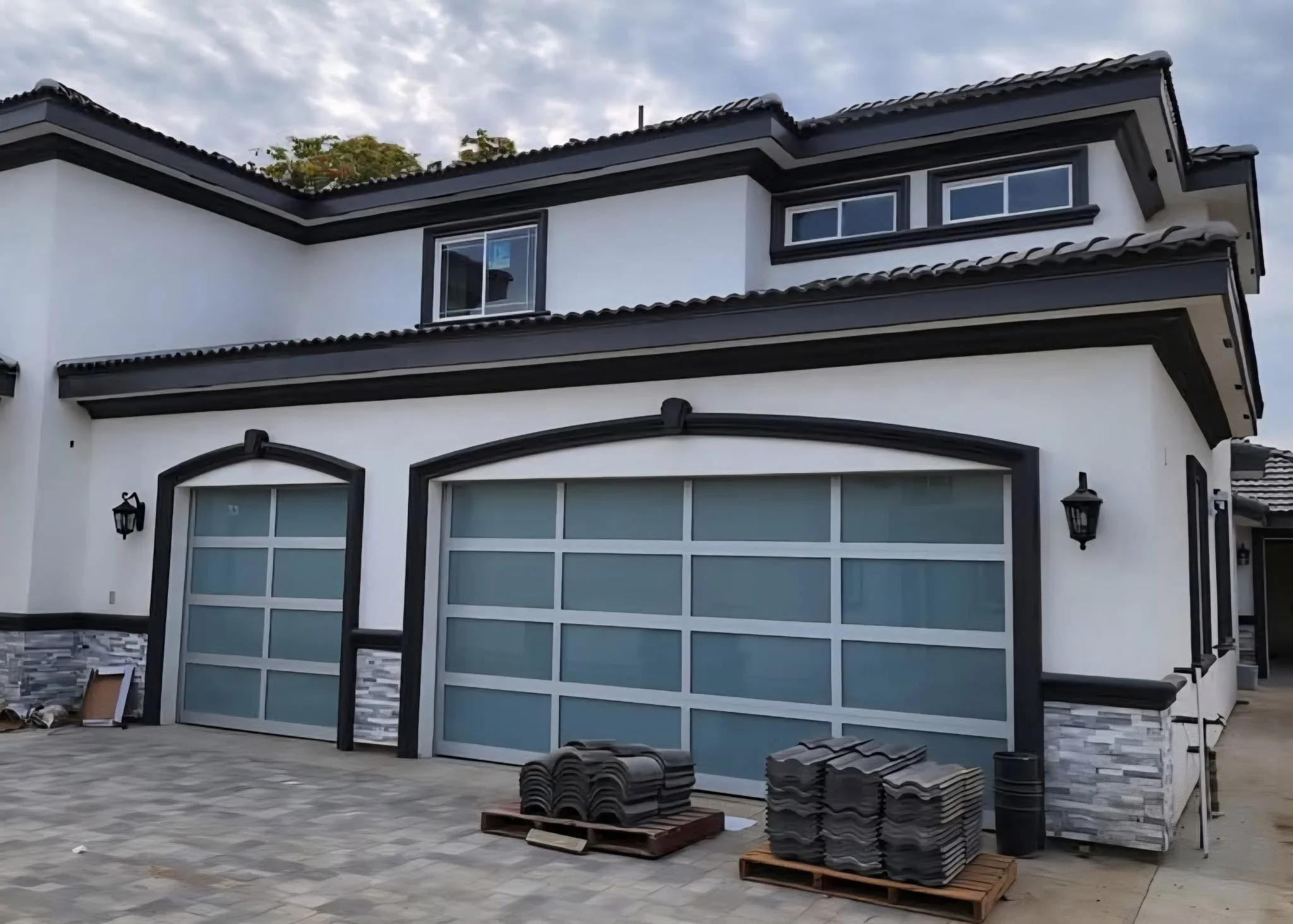 Modern two-story house with white walls and black trim. Two garage doors and exterior wall-mounted lamps. Construction materials, tiles, and pallets in front.