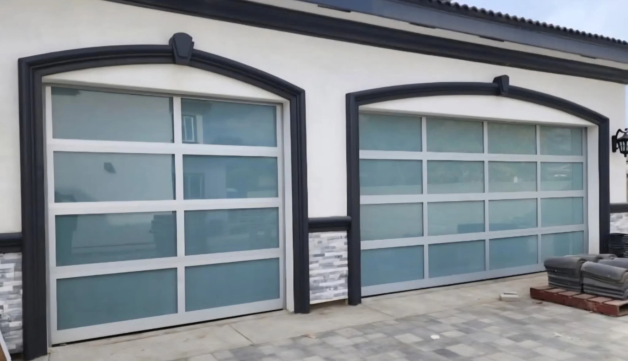 Two modern garage doors with frosted glass panels, black framing, and decorative trim, set in a white house with stone accents and a paved driveway.
