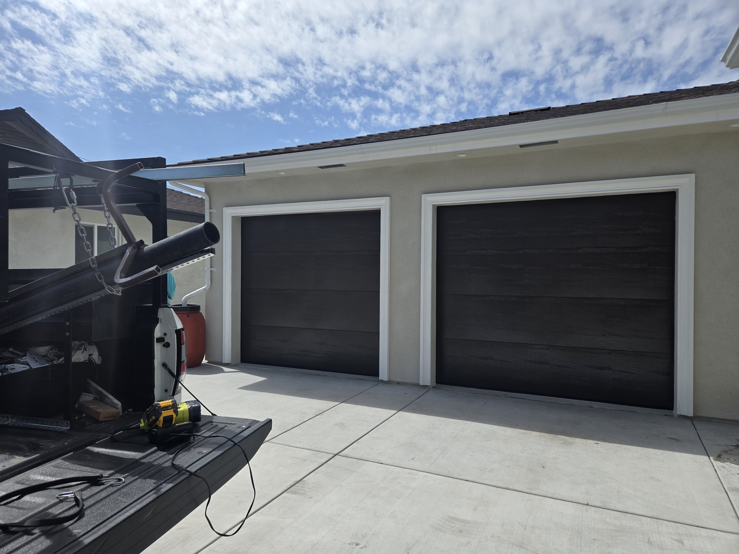 A residential garage with two black garage doors and a concrete driveway. There are some tools and equipment on a work surface in the foreground, including a yellow and black cordless drill. Part of a patio or outdoor area with a barbecue grill and a