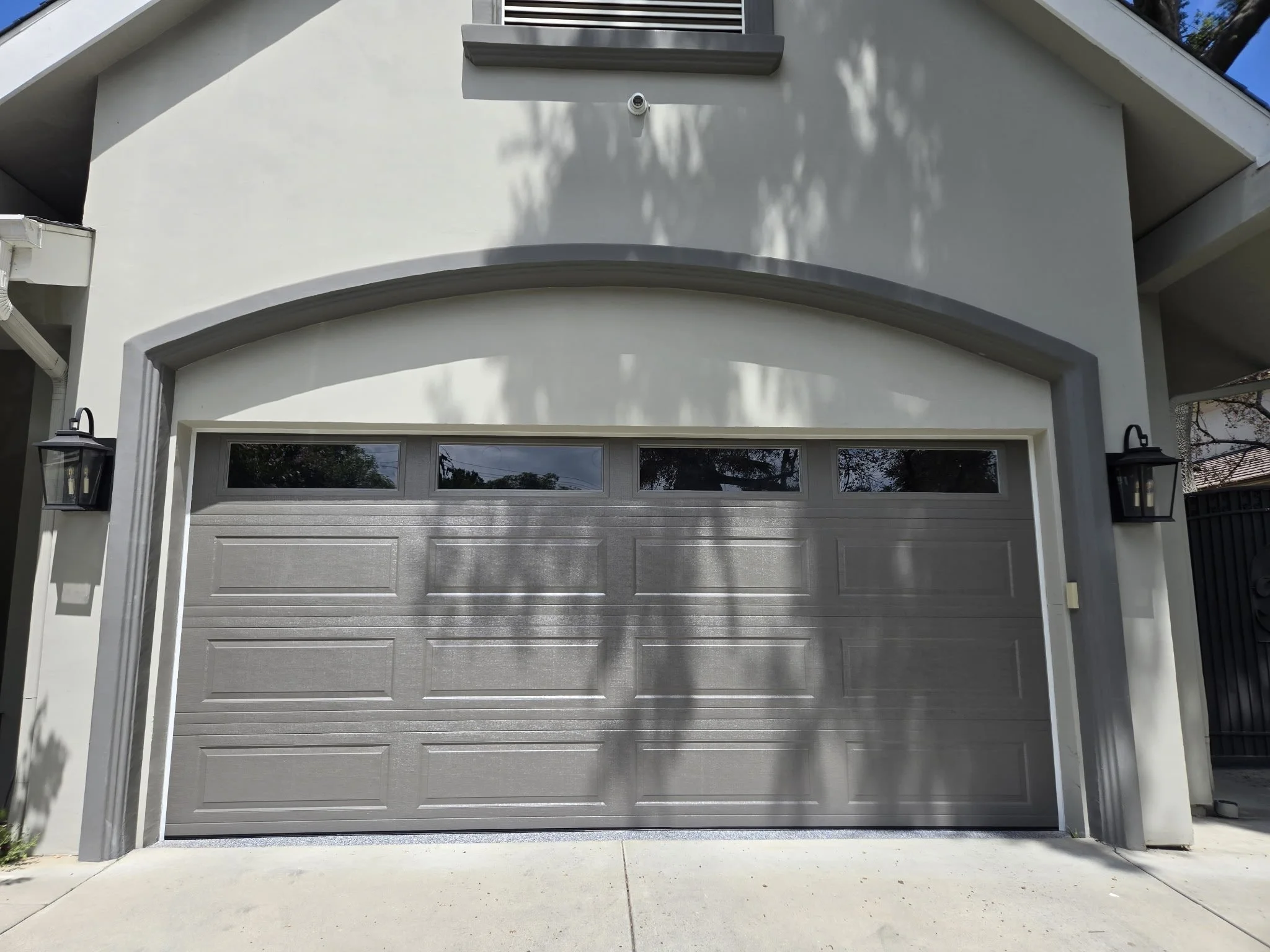 Gray garage door with four small windows at the top, flanked by two black wall-mounted lanterns, on a white house with shadows of trees on the facade.