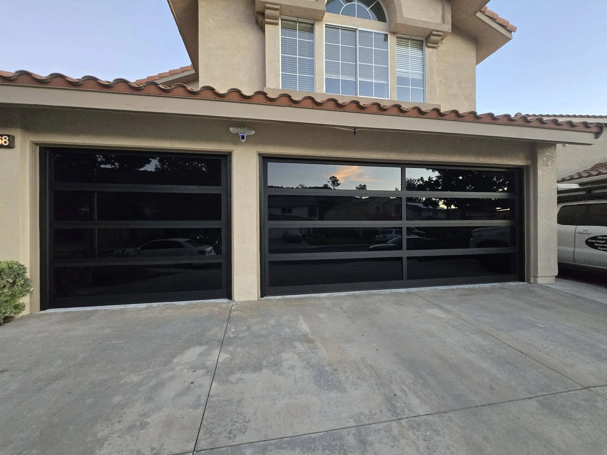 Front view of a two-car garage with black glass panel doors, part of a beige multi-story house with a tiled roof, and a concrete driveway in the foreground.