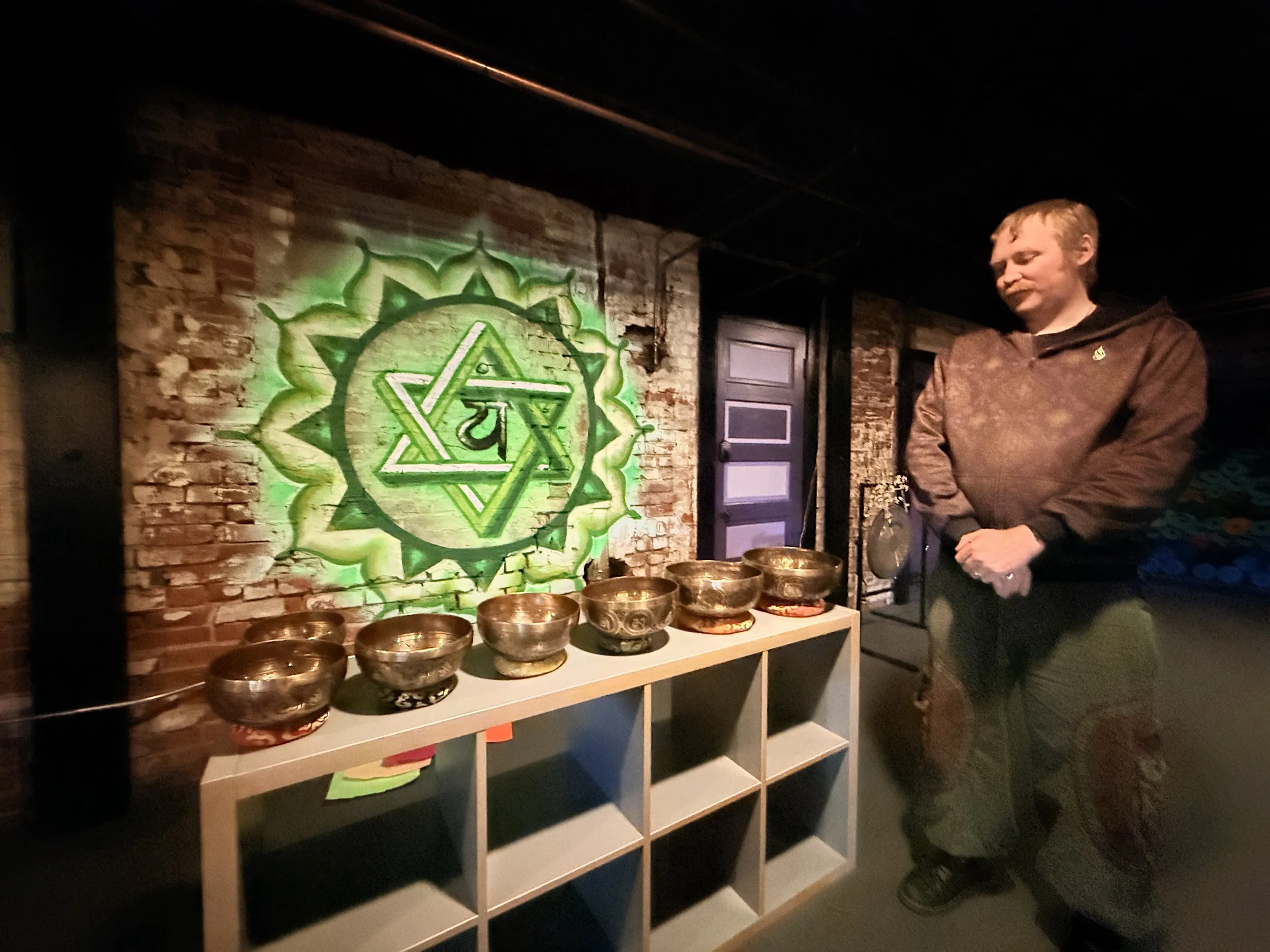 A man stands indoors with a row of singing bowls on a white shelf in front of a brick wall painted with a green symbol featuring a star of David and a central circle with a design inside.