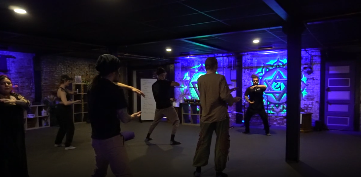 People practicing a choreographed dance or martial arts move in a dimly lit room with blue neon lighting and exposed brick walls