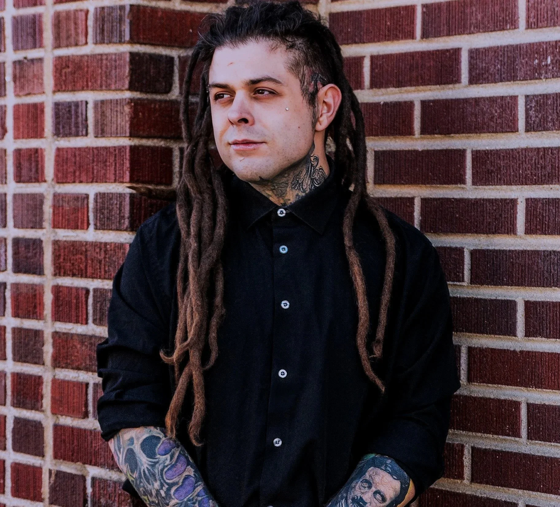 A man with long dreadlocks, tattoos on his arms and neck, wearing a black shirt, standing against a brick wall.