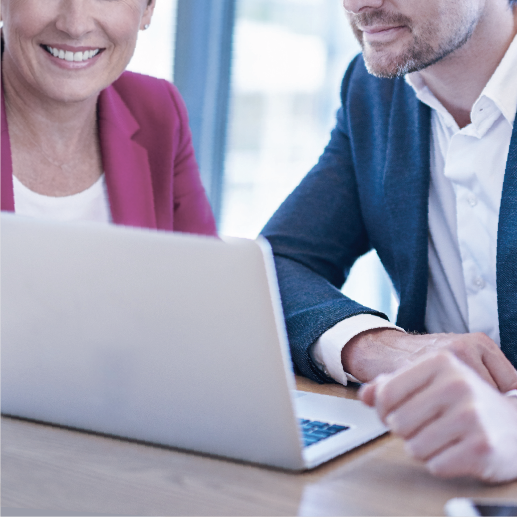 Two business professionals, a woman in a pink blazer and a man in a navy blazer, smiling and working together on a laptop in a modern office setting.
