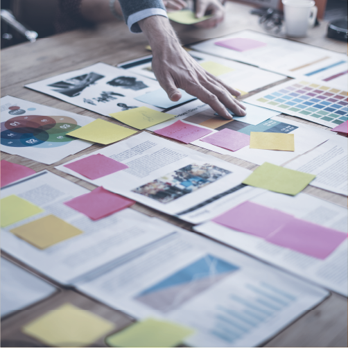 A person's hand reaches over a cluttered wooden table filled with papers, printed images, color swatches, and colorful sticky notes, suggesting a brainstorming or planning session.