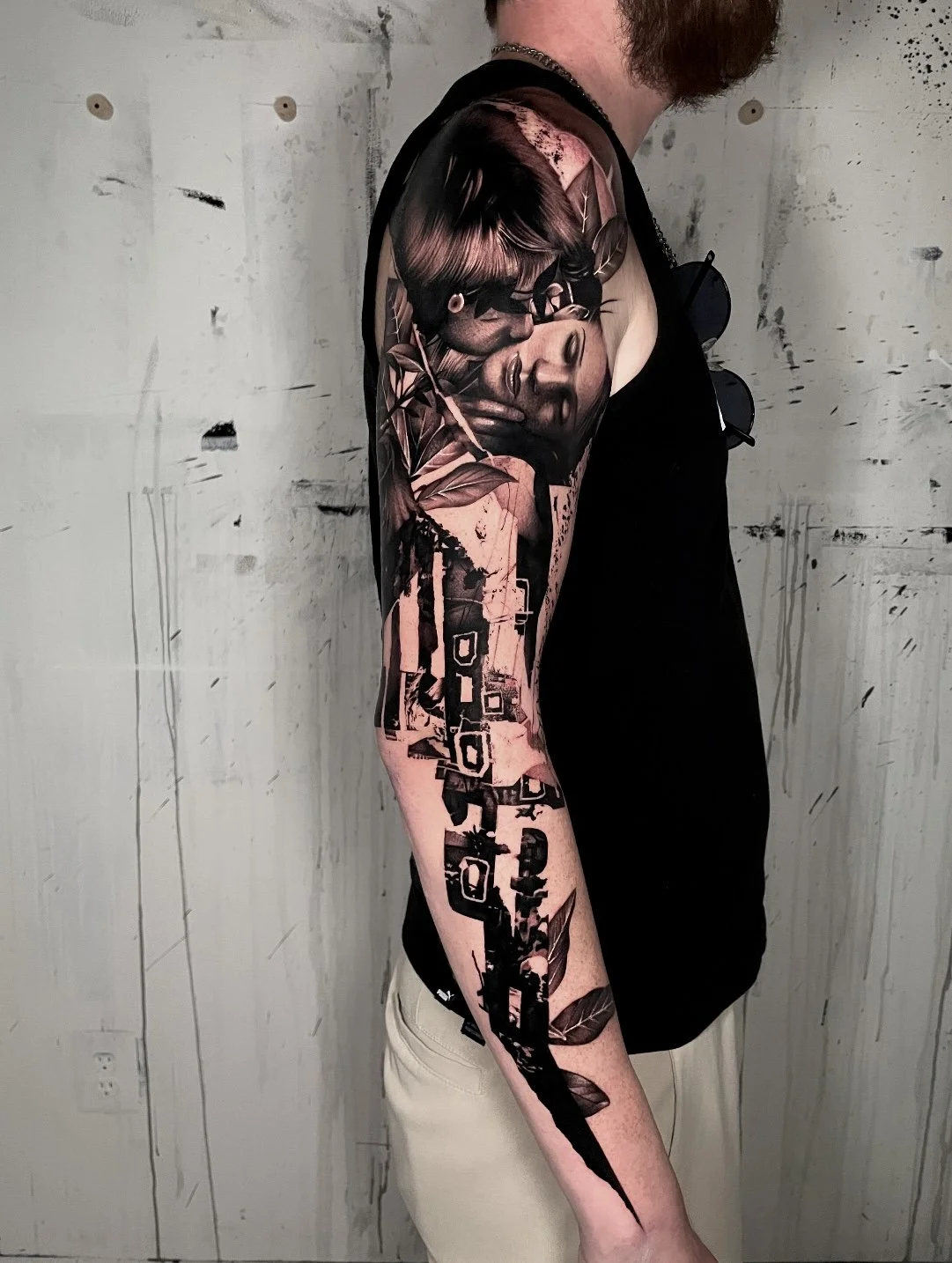 A person with a large, detailed tattoo on their left arm, depicting a woman's face surrounded by leaves and abstract elements. The person is wearing a sleeveless black top and beige pants, standing against a textured gray wall.