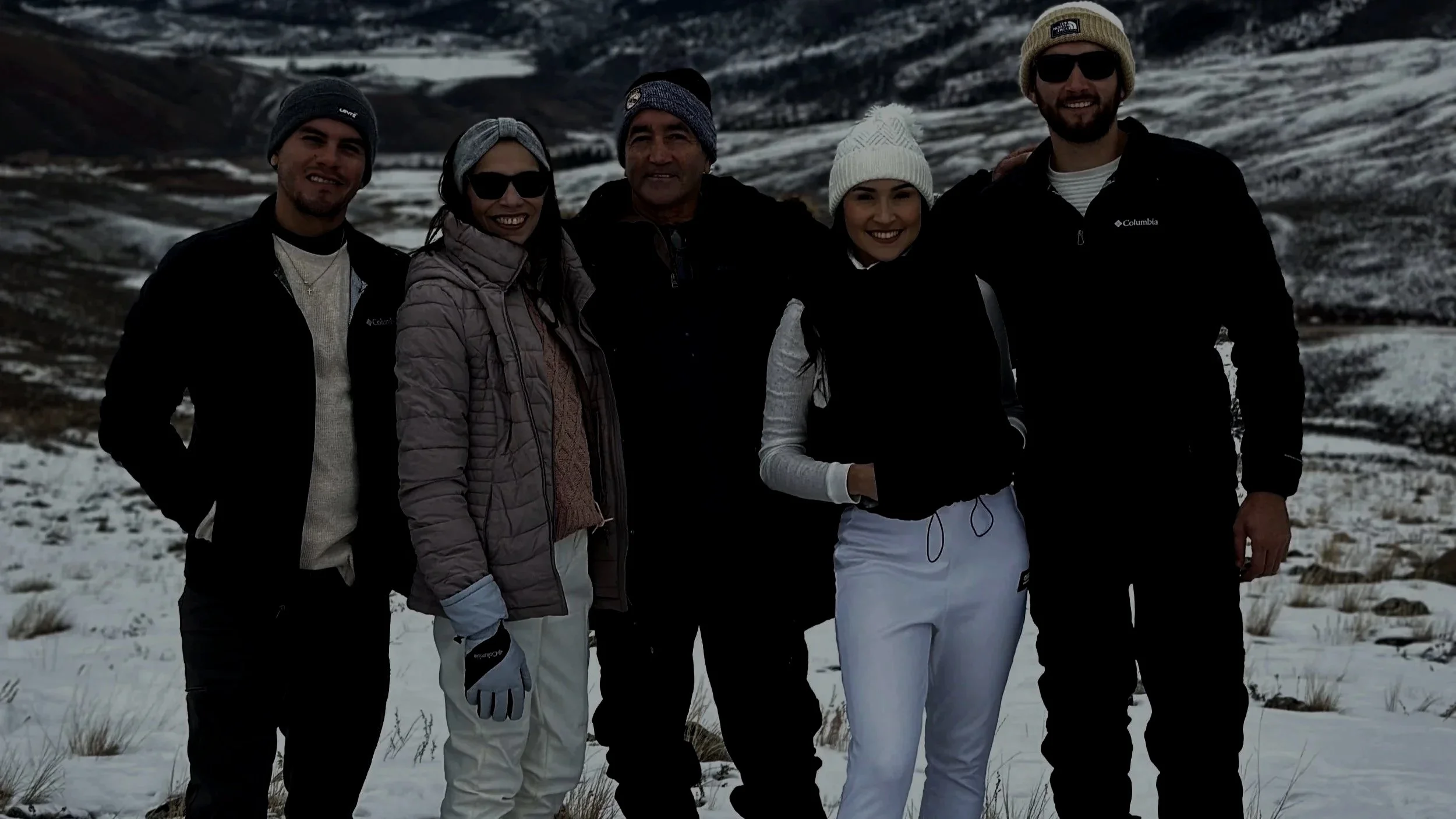 Group of six people standing outside in snowy landscape, dressed in winter clothing, smiling at the camera.