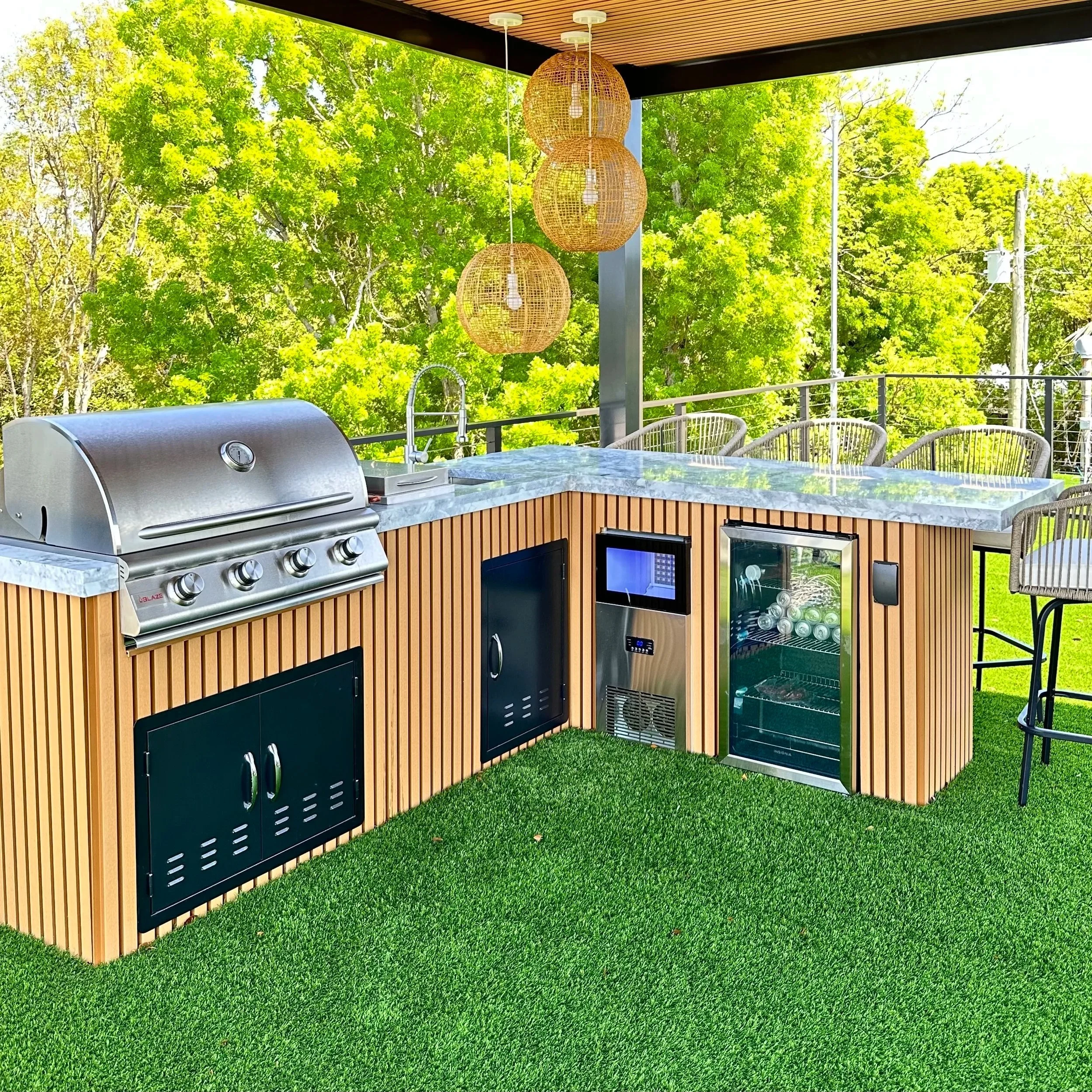 Outdoor kitchen with stainless steel grill, mini fridge, and microwave built into a wooden counter with marble top, set on artificial grass with green trees in the background. Hanging wicker lamps are above, and there are bar chairs around the counter.
