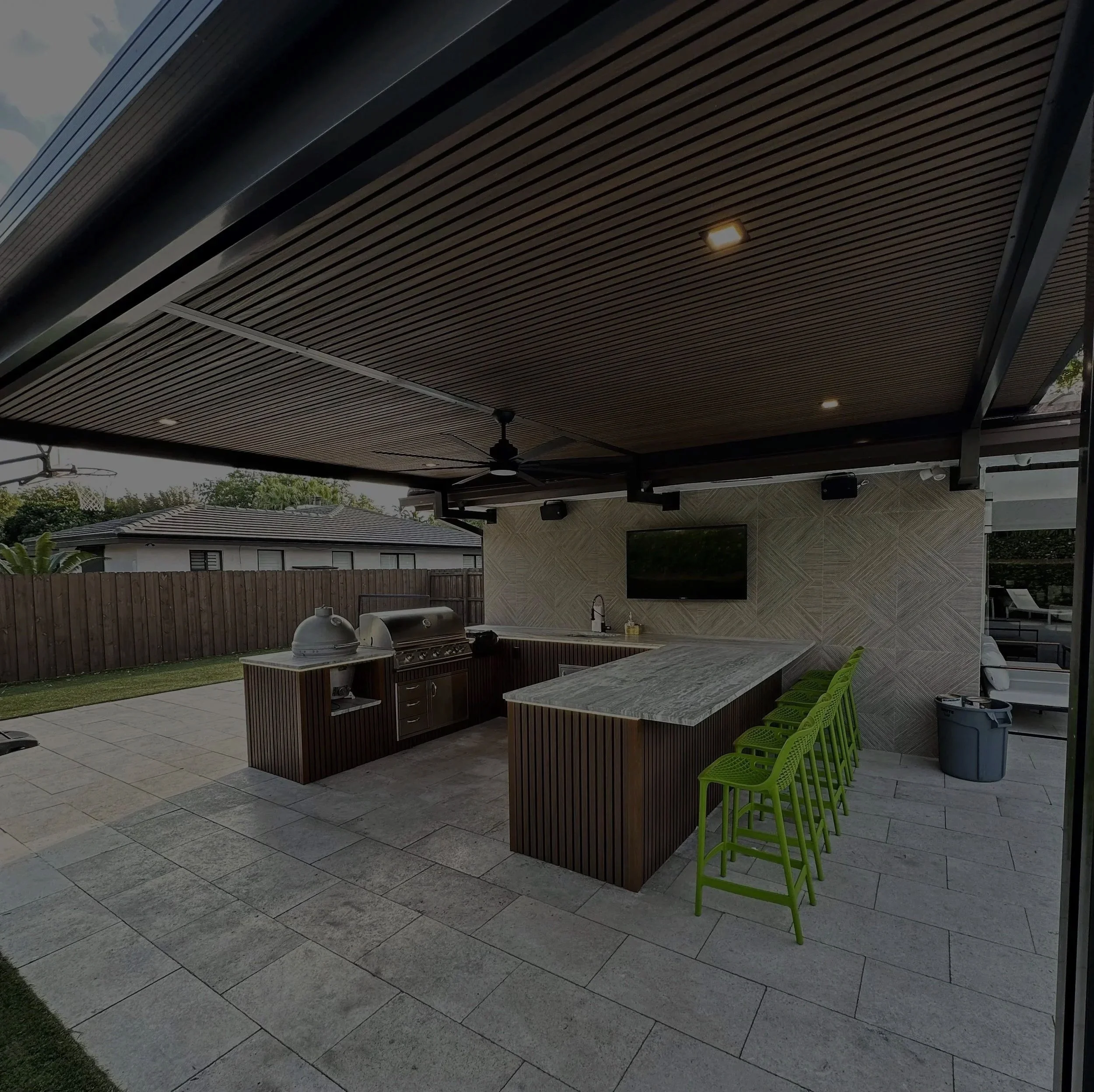 Outdoor kitchen with a bar, green chairs, grill, TV, ceiling fan, and shaded roof.