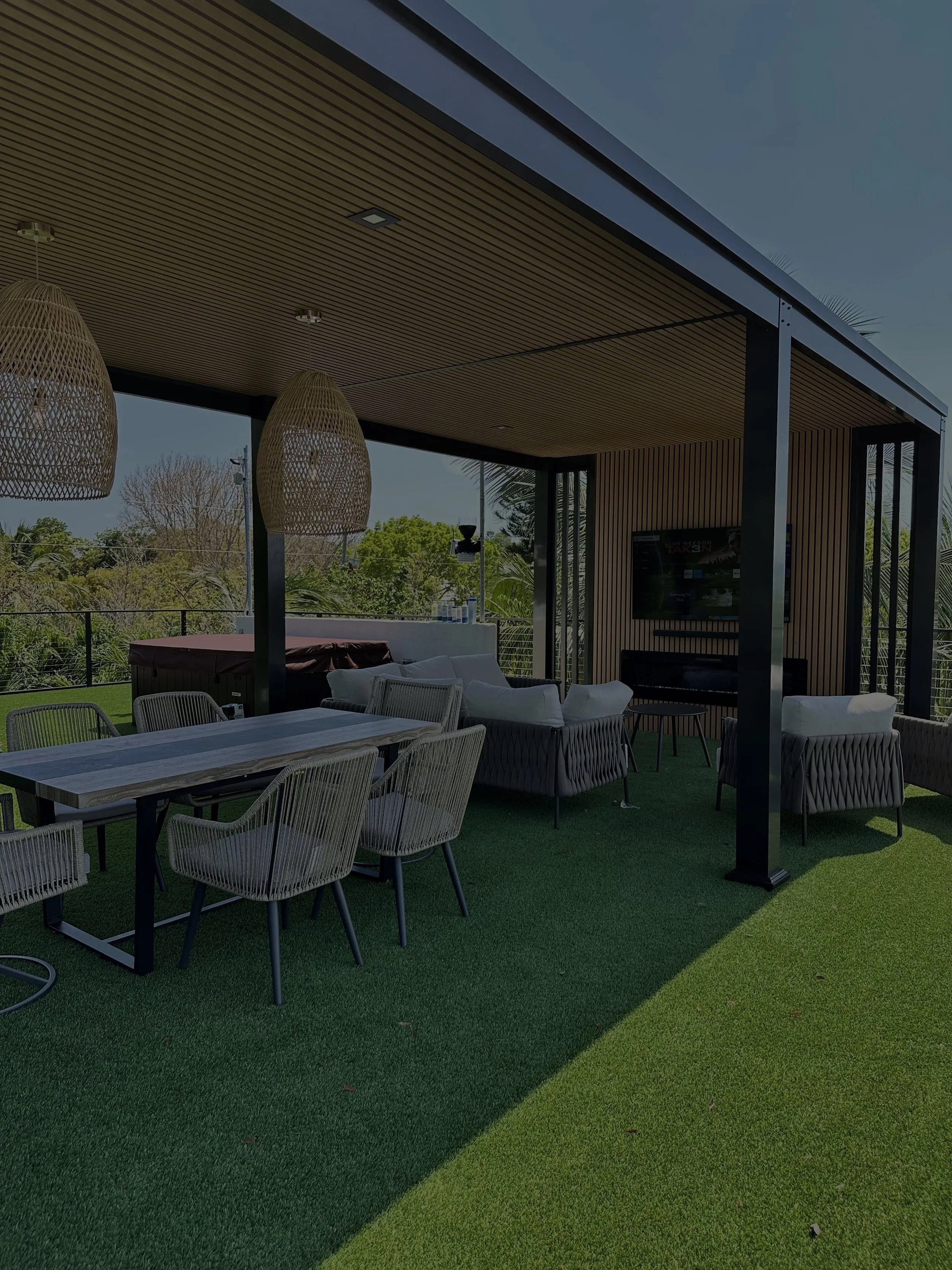 Outdoor patio with dining table and chairs, couches, hanging wicker lights, television, and a view of trees and greenery.