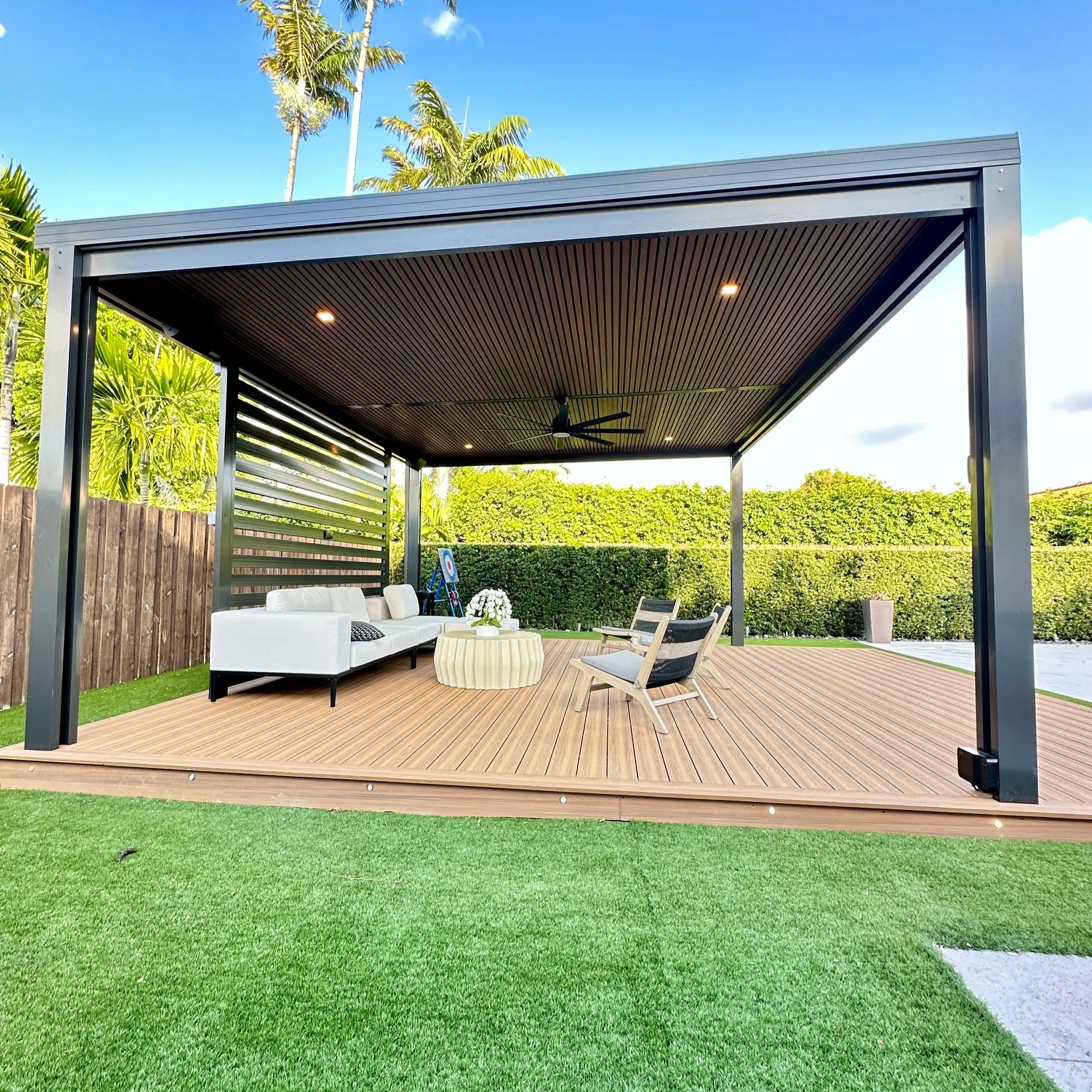 An outdoor patio area with wooden decking, a black metal-framed roof, ceiling fan, white sofa, two wooden chairs, a round table with flowers, and lush green hedges and palm trees in the background.