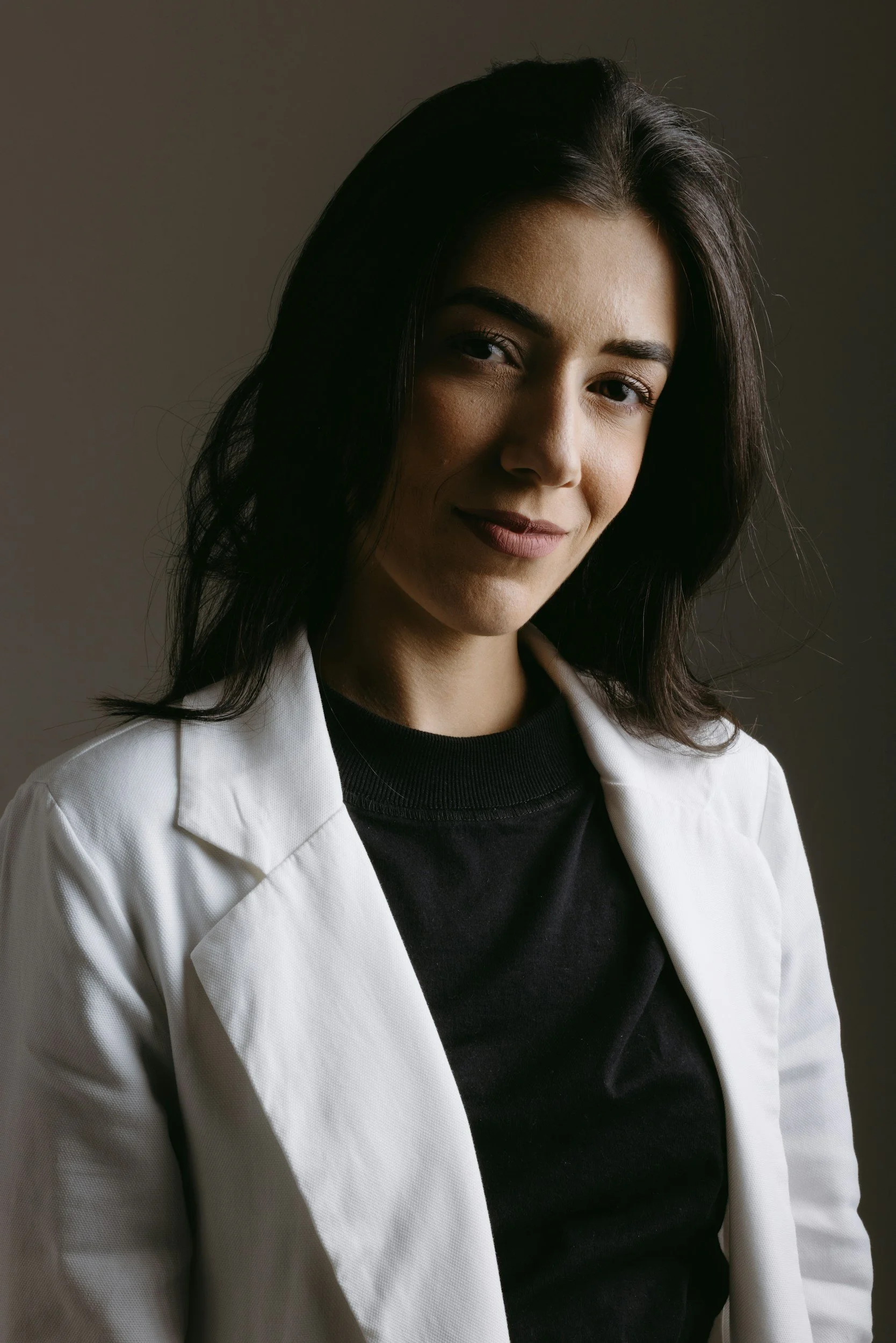 A woman with dark hair and light skin, wearing a white blazer over a black top, smiling softly against a dark background.