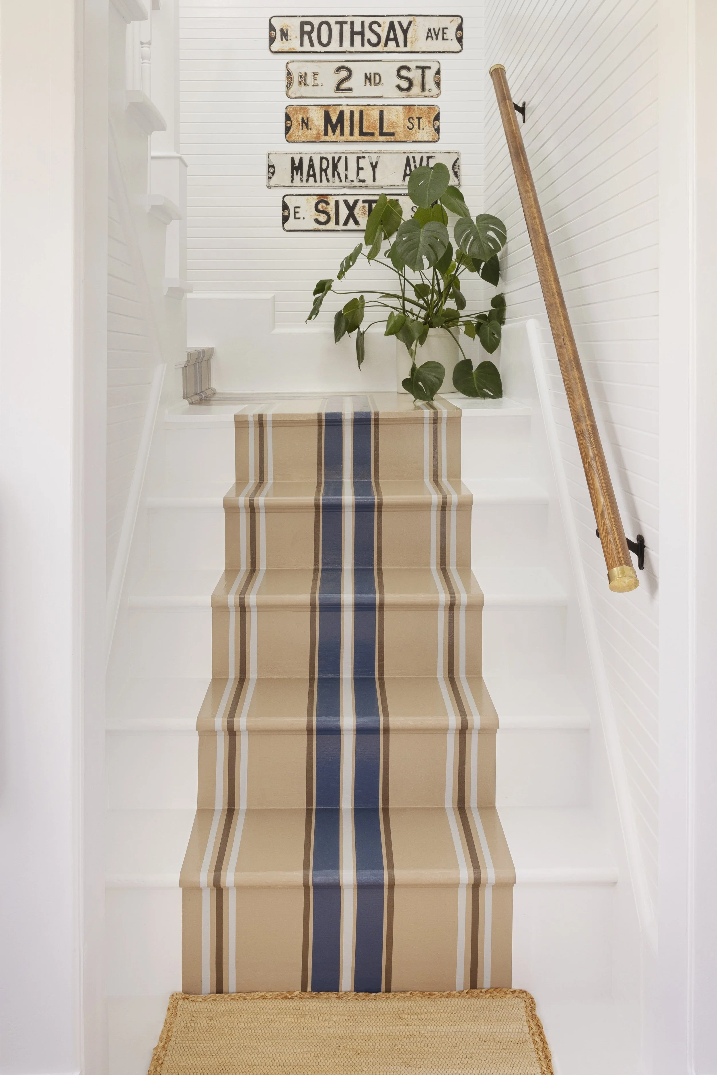 A staircase with a beige and blue striped runner, a potted plant with large green leaves, and vintage street signs on the wall at the top of the stairs.