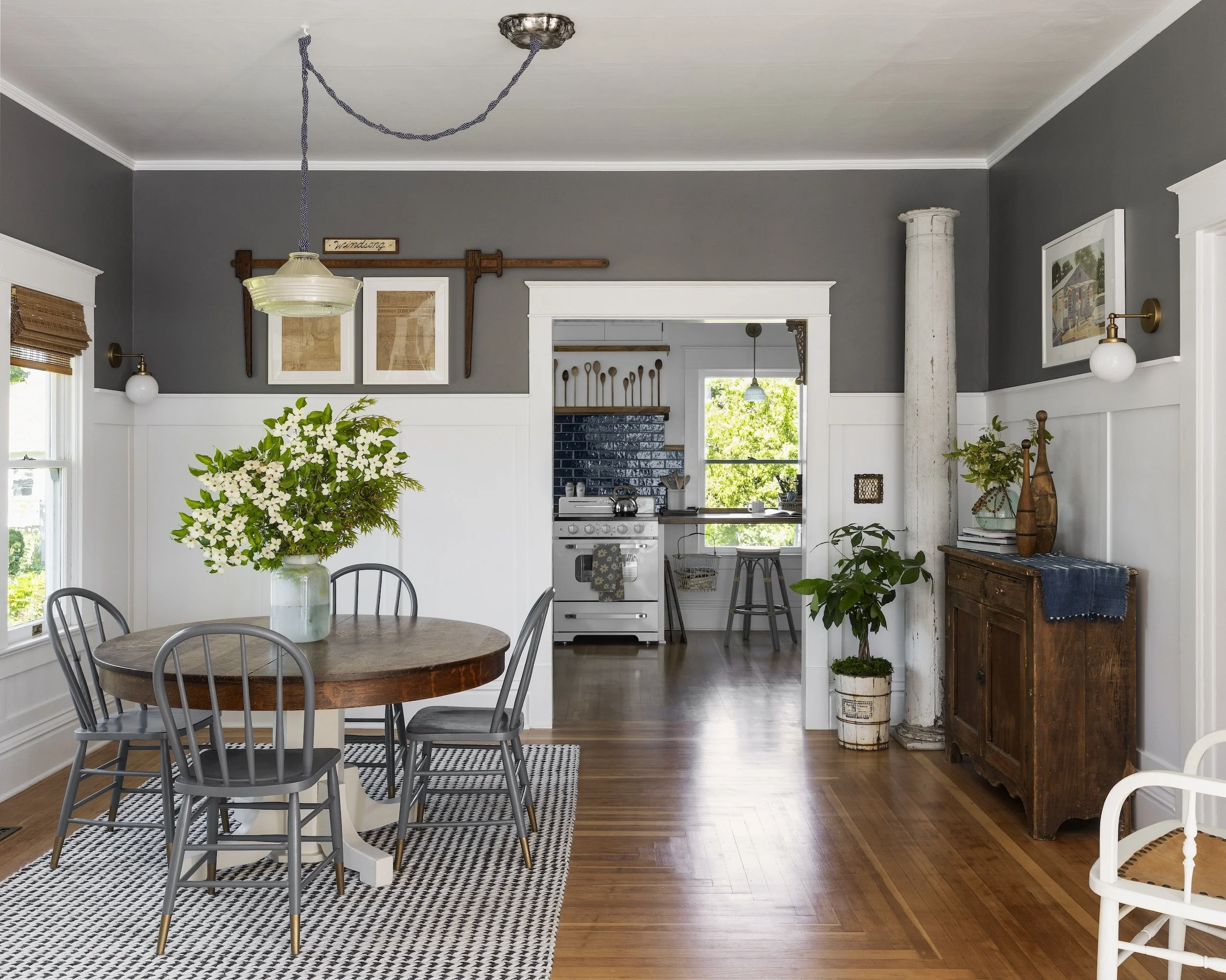 A dining room with a round wooden table, four gray chairs, a vase with white flowers, and a black and white patterned rug. The room has white wainscoting, gray walls, and wooden flooring. There is a doorway leading to a kitchen with a white stove, bl