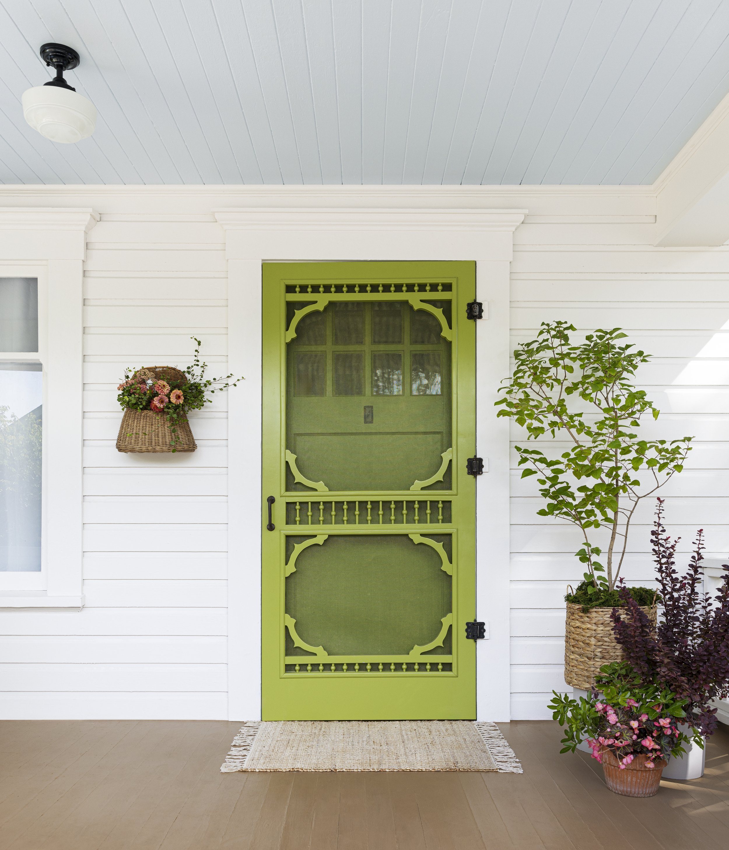 Green decorative screen door with white house siding, potted plant with green leaves, wall basket of flowers, and front porch rug.