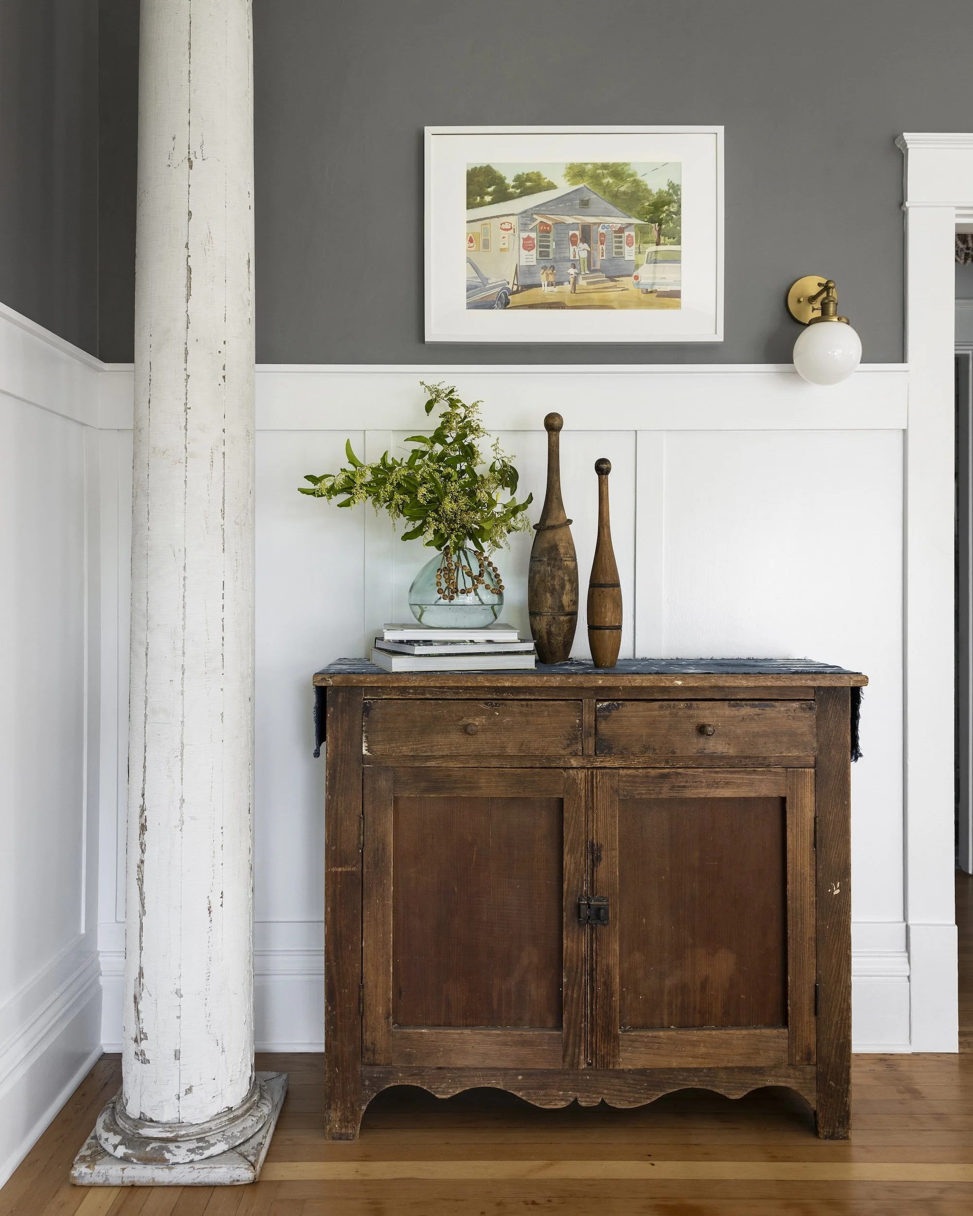 A vintage wooden sideboard with a bouquet of greenery and decorative wooden vases on top, set against a half-white, half-gray wall with framed artwork, a white wall sconce, and a white-painted wooden column nearby.